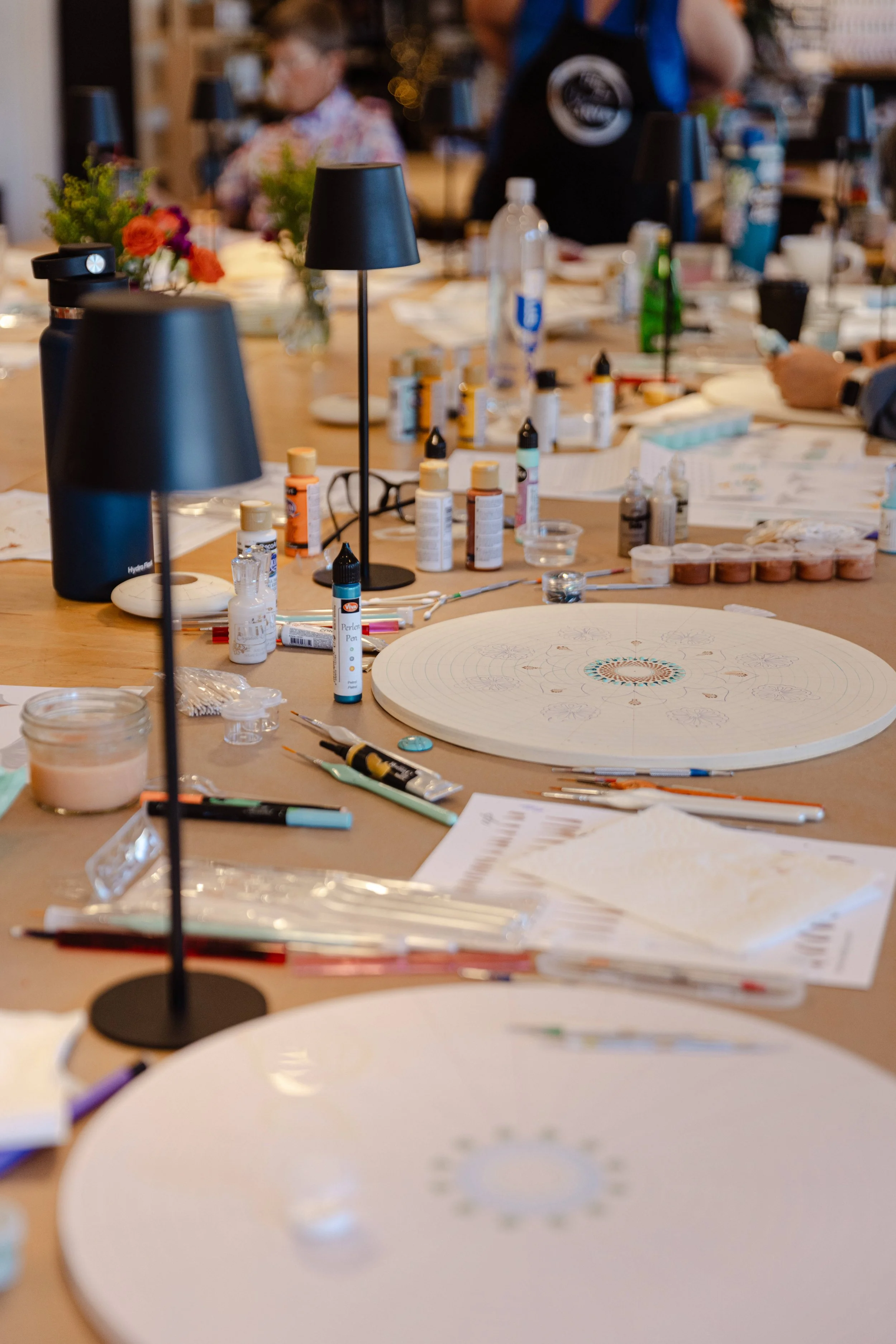A workspace with various art supplies, including paint bottles, brushes, and a round canvas with sketches, surrounded by lamps and papers. In the background, people are gathered around the table.