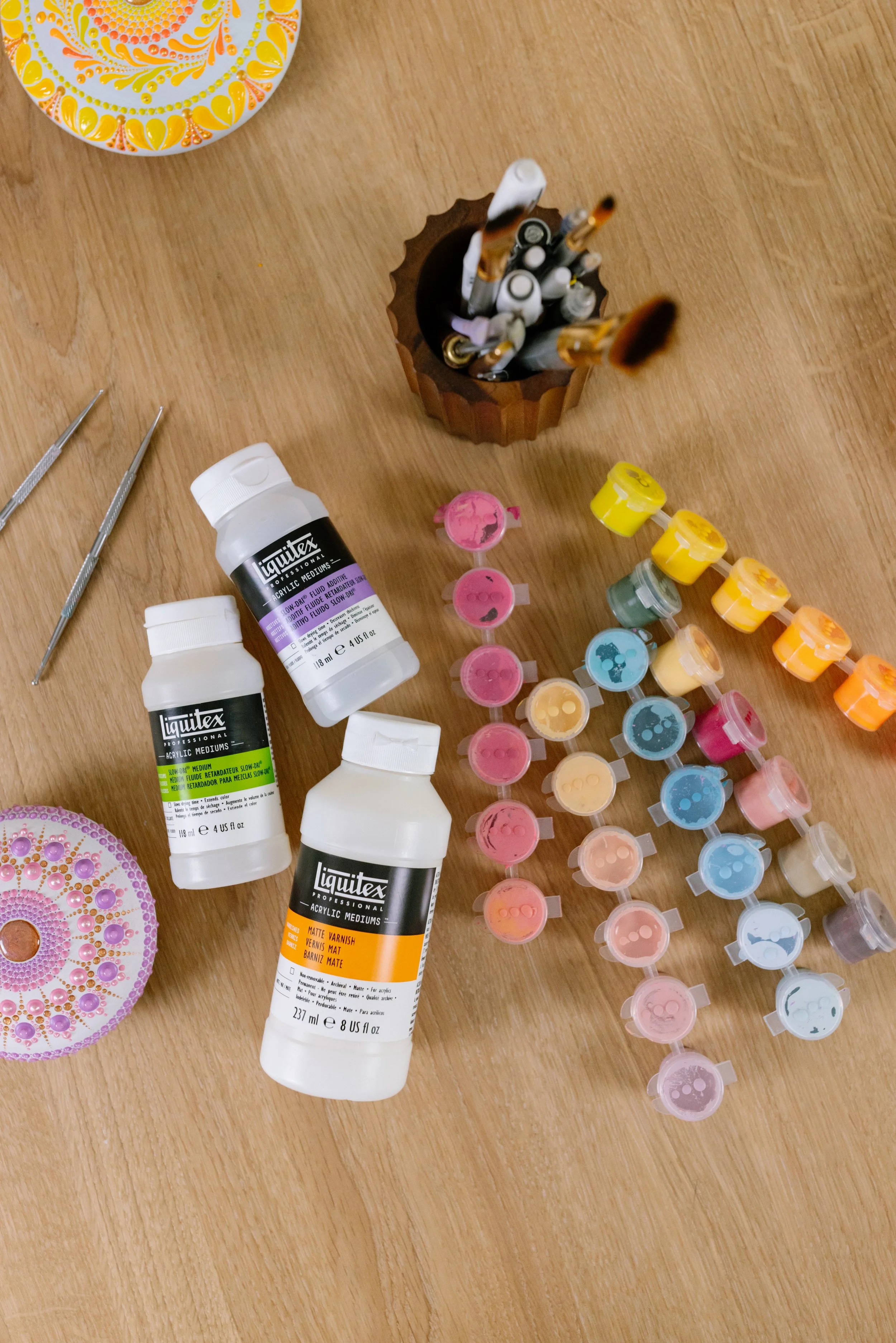 An overhead view of nail art supplies on a wooden surface, including bottles of Liquitex acrylic medium, a color palette, paint swatches, and a container with paintbrushes.