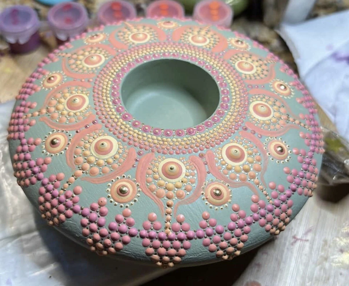 Decorative ceramic bowl with pink and peach dot patterns and spiral designs, resting on a surface with paint supplies nearby.
