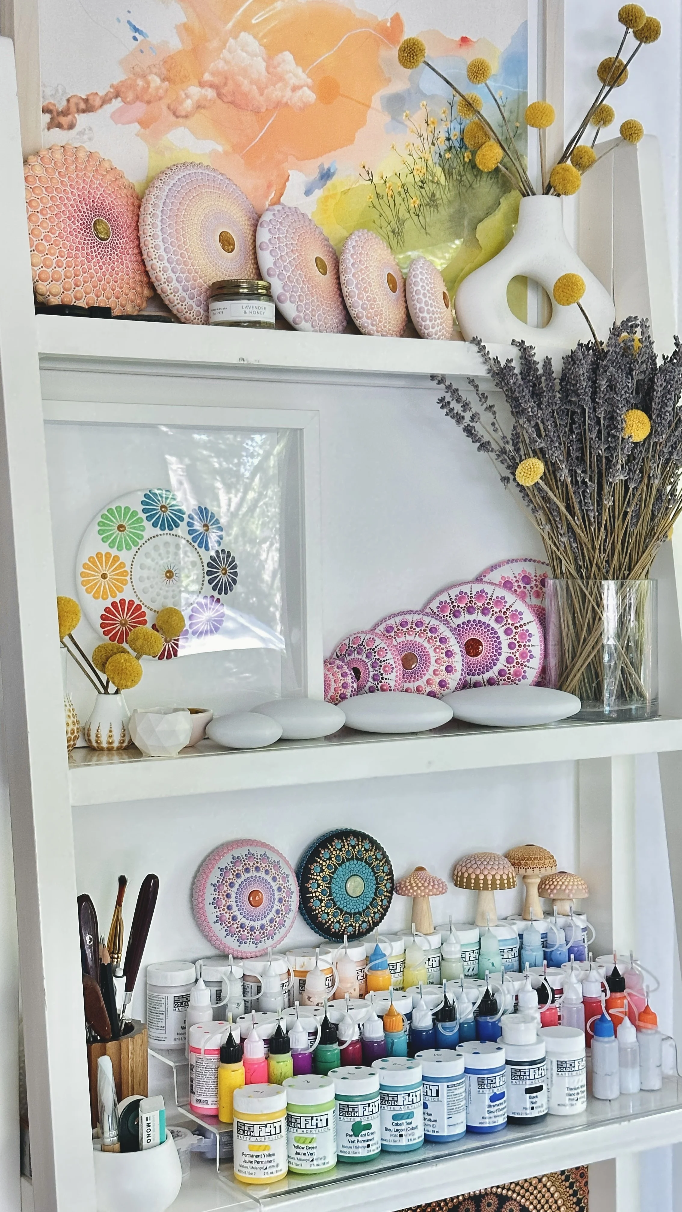 Display of decorative art pieces including circular designs, floral artwork, ceramic mushrooms, and bottles of acrylic paint on a white shelf with vases and dried flowers.