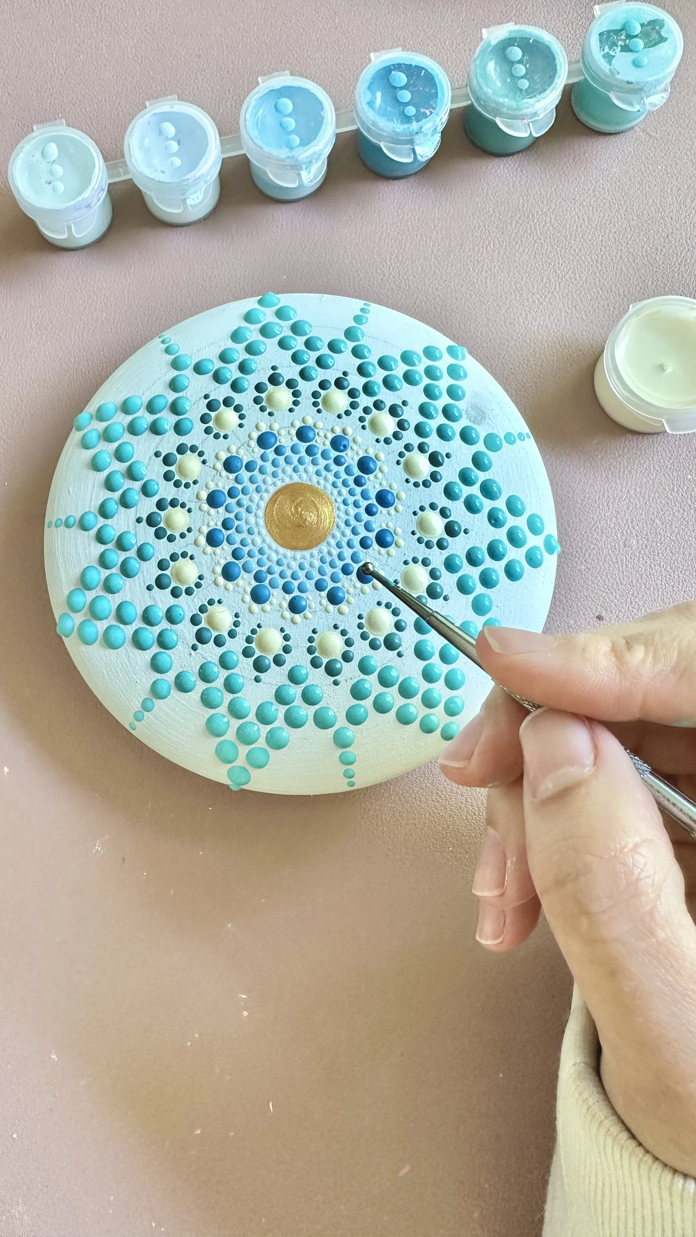 A person creating a dot mandala art on a round white surface using small blue and white paint dots and a dotting tool. Several small containers of blue and white paint are visible above the art, and the background is a light pink surface.