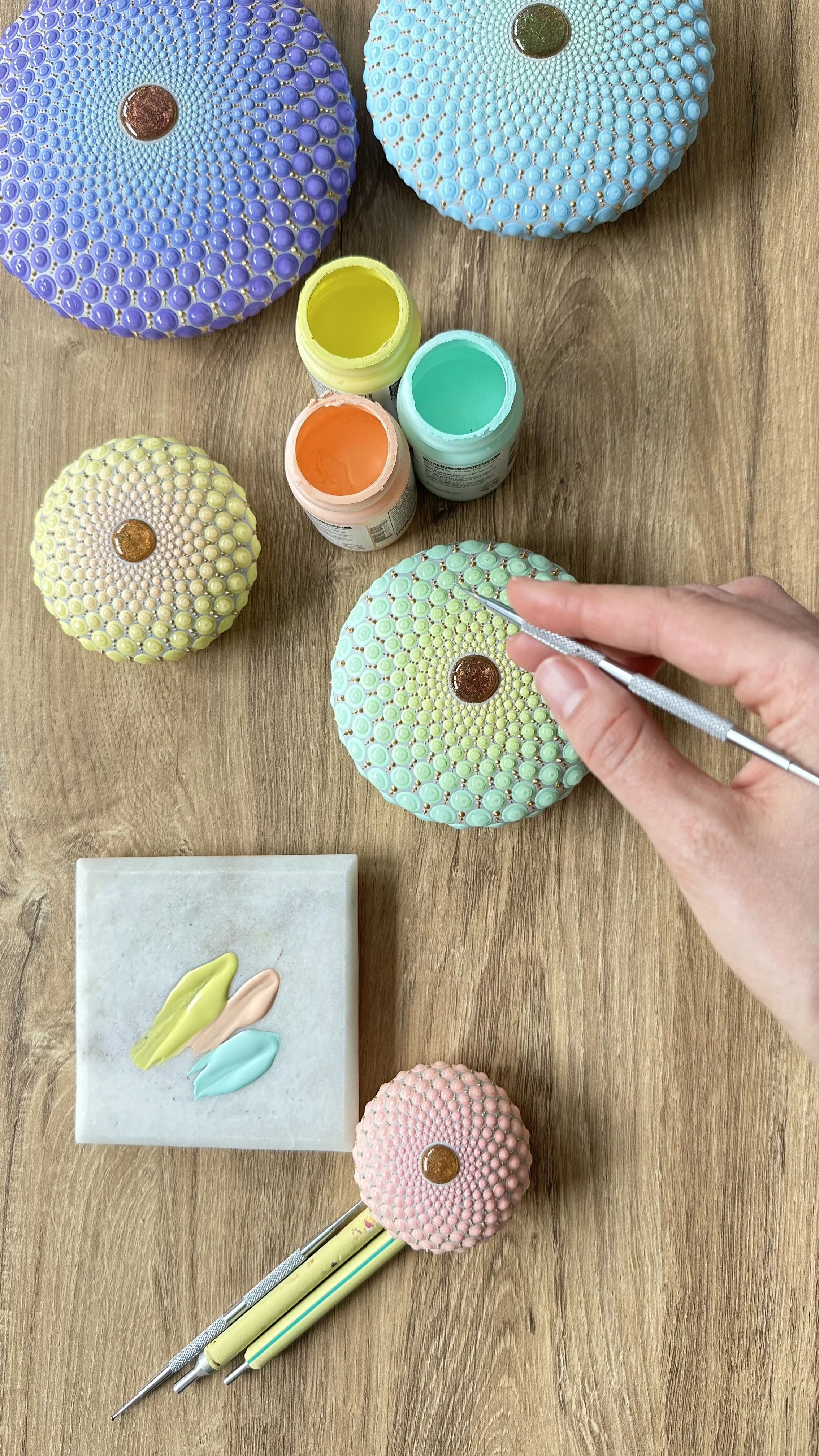 Person painting colorful textured ceramic objects with dot patterns using fine tools, surrounded by jars of paint and additional ceramic pieces on a wooden surface.