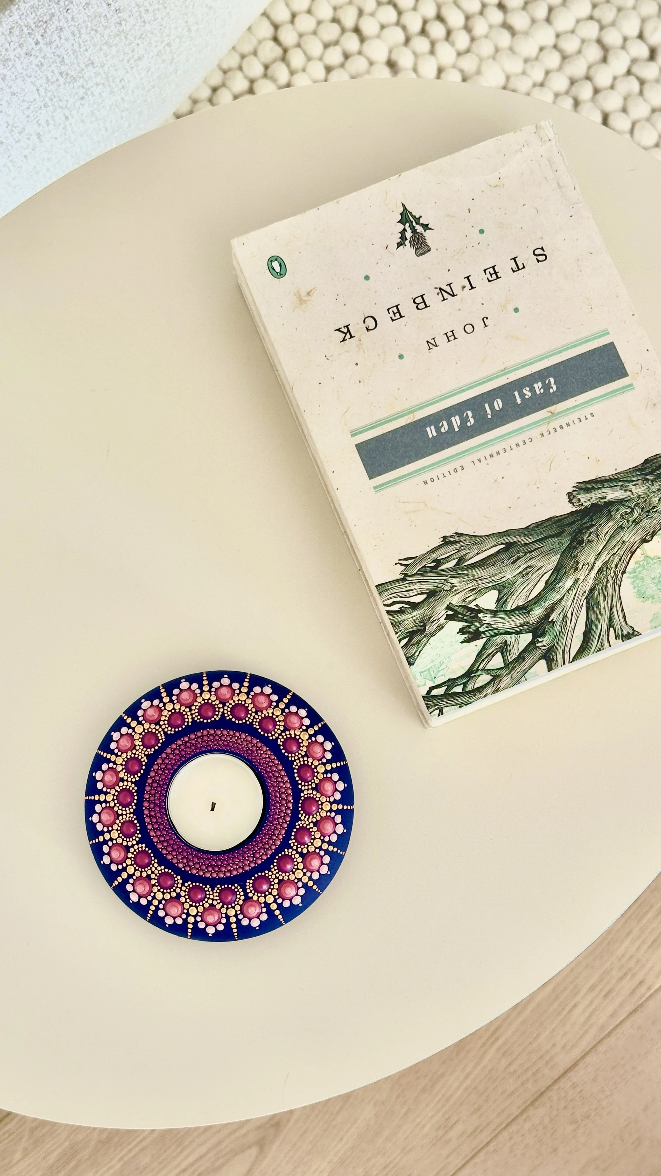 A white round table with a colorful circular candle holder and a book titled 'Steinbeck' placed on it, with a textured wall in the background.