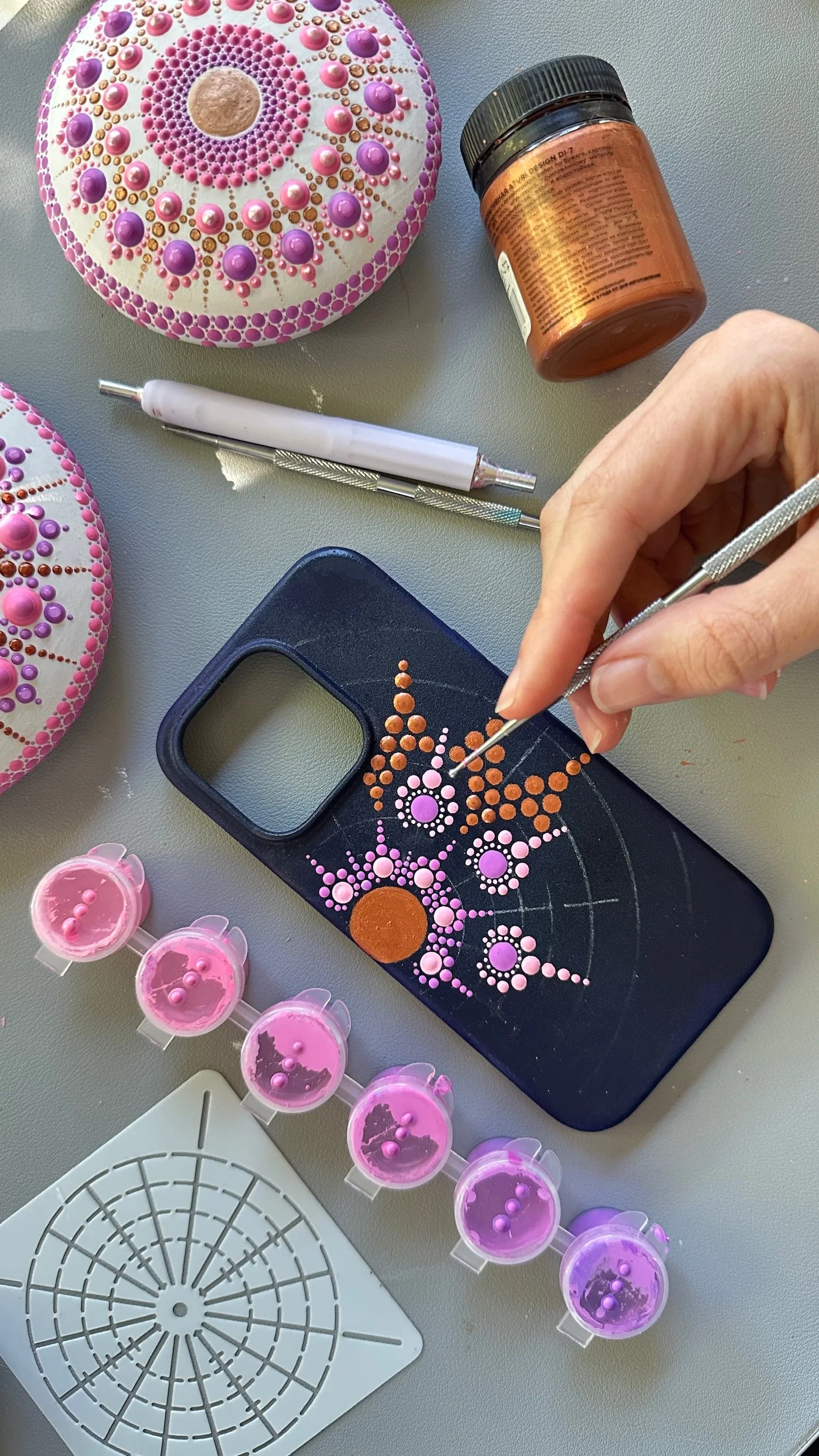 A person creating a dot art design on a phone case using small paint dots, with various tools and paints around.