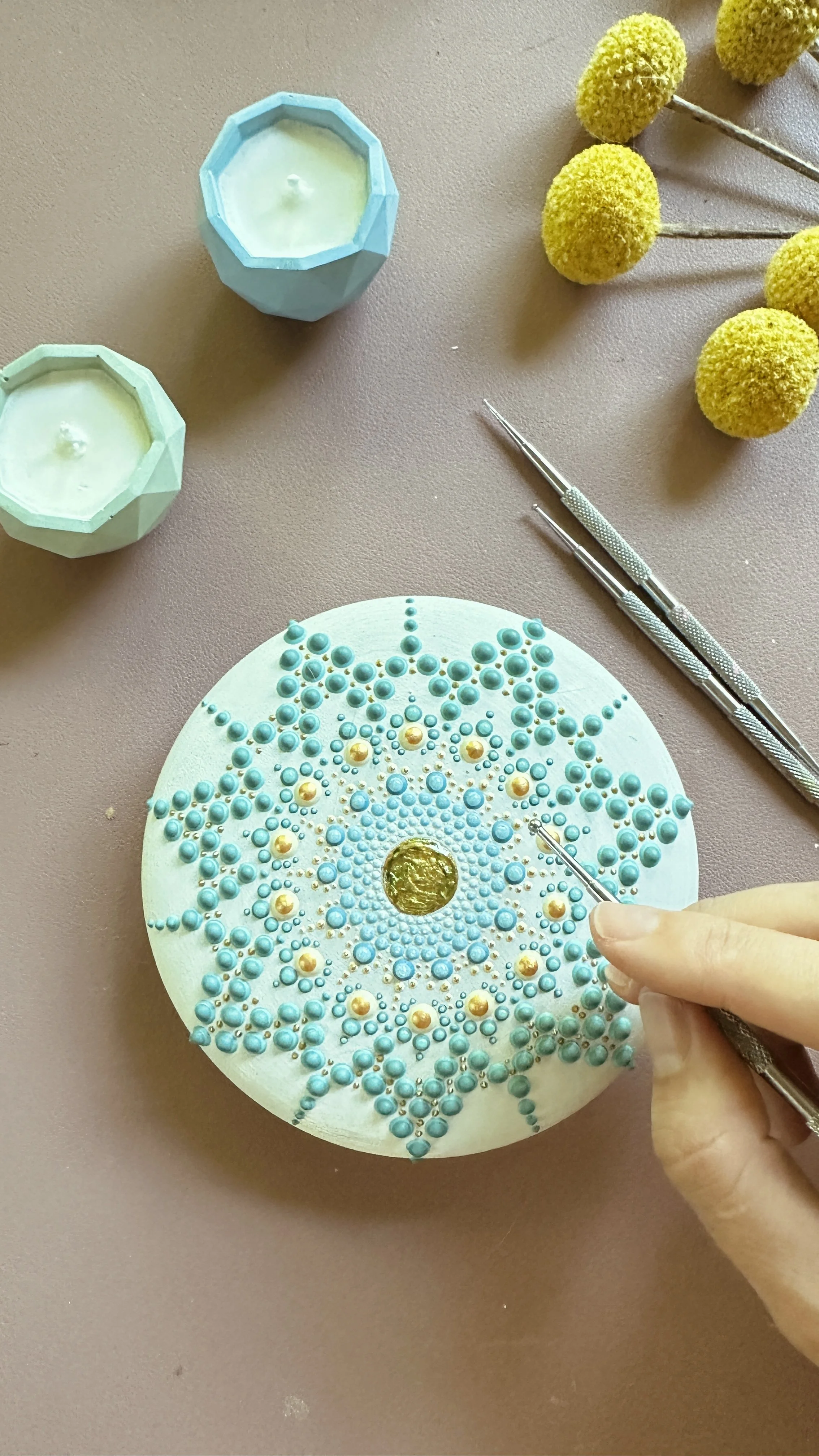 A person decorating a circular object with small blue and gold beads to create a detailed design. There are also yellow flowers, candles, and craft tools on the table.