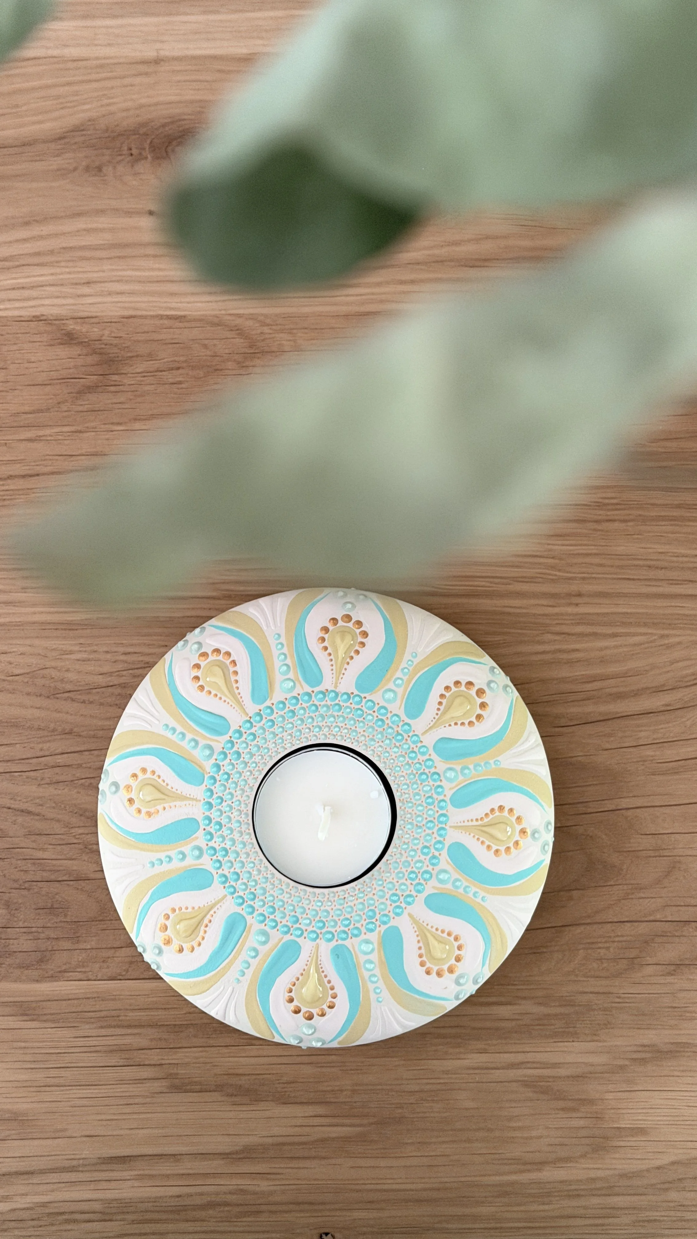 A decorative candle holder with intricate turquoise, beige, and gold patterns, holding a white candle on a wooden surface, with a blurred green plant leaf in the foreground.