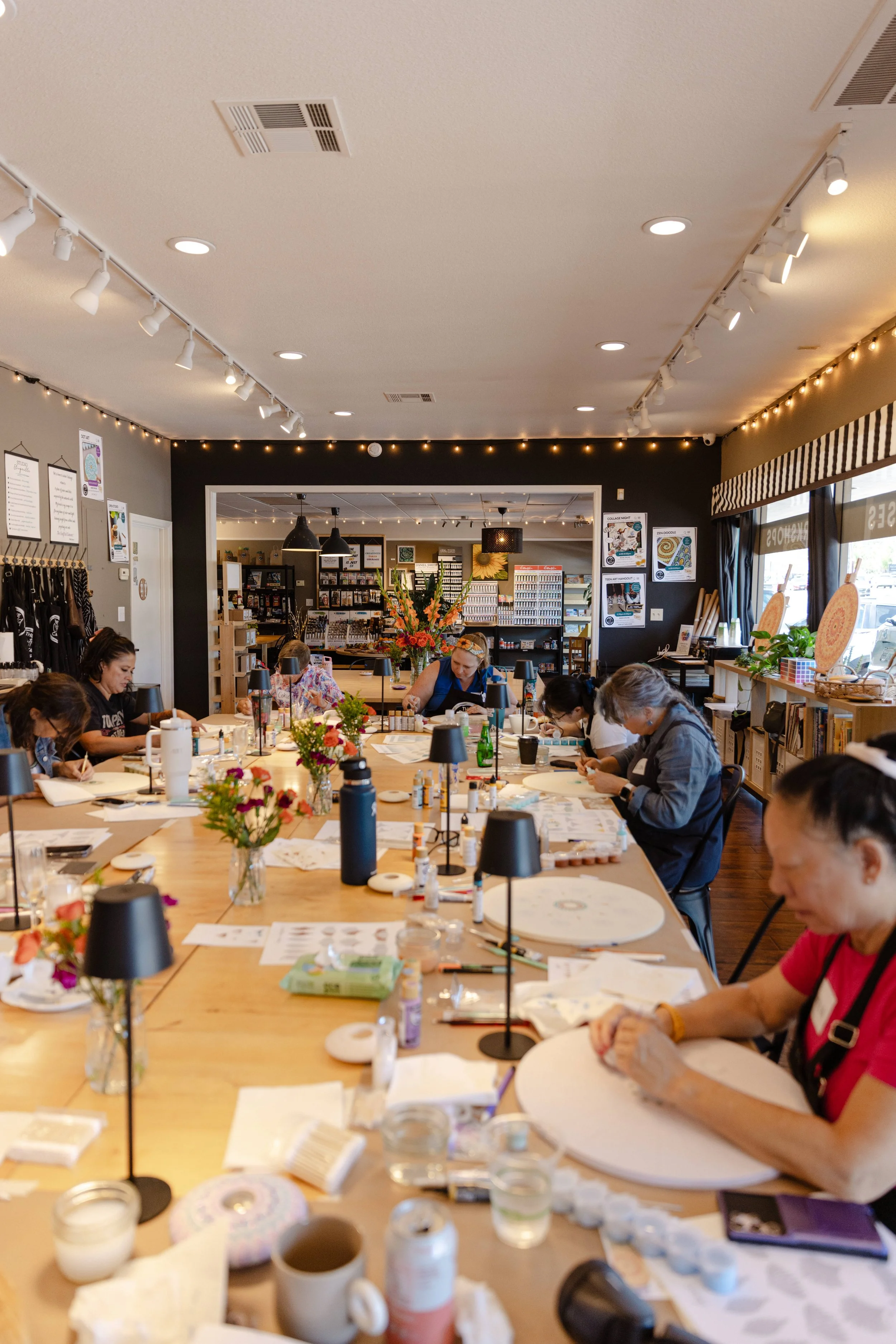 A group of people sitting around a large wooden table engaged in a craft activity, with art supplies, flower arrangements, and various decorative items on the tables in a well-lit room with string lights and posters on the walls.
