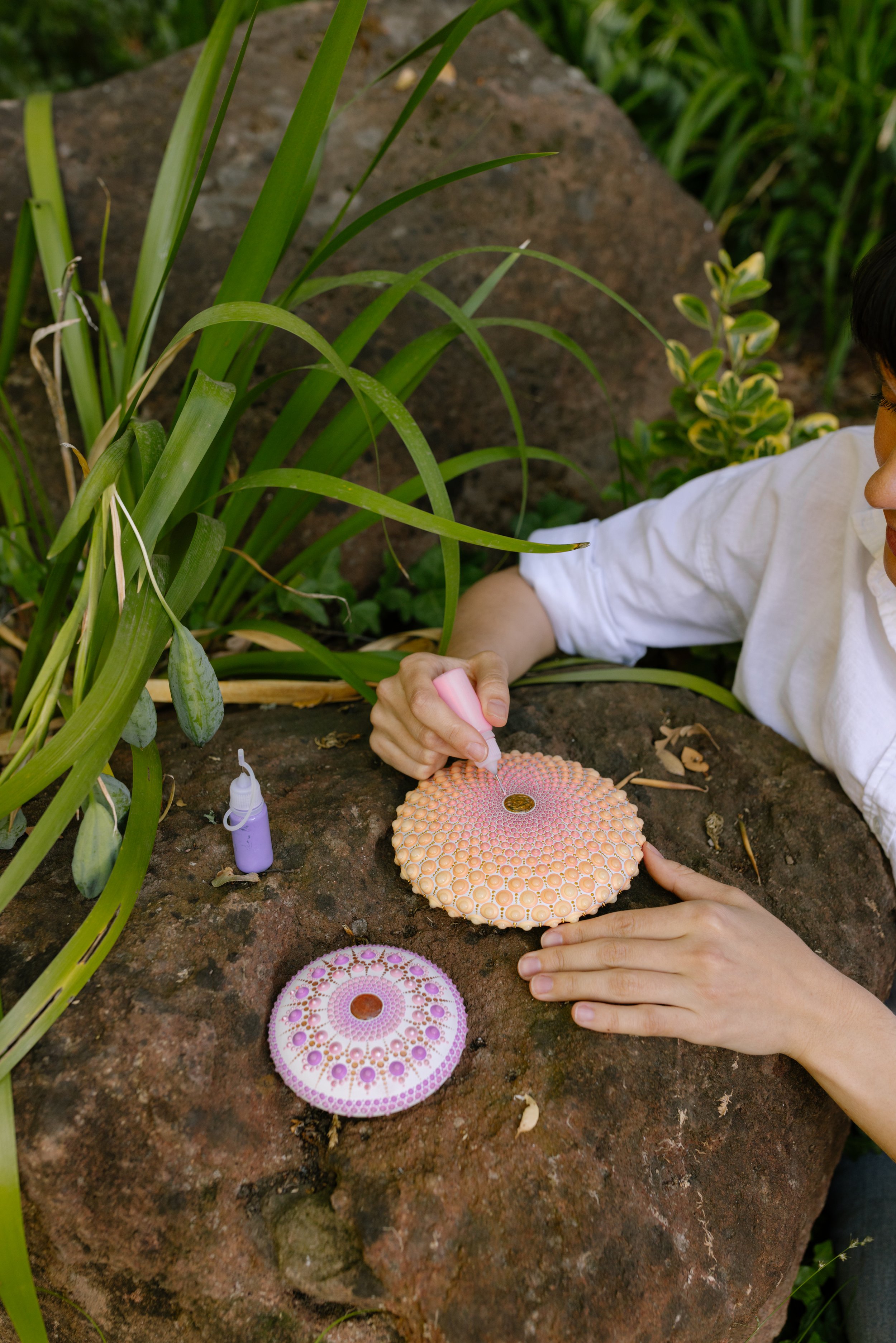A person in a white shirt creates dot paintings on circular canvas using small tools in an outdoor garden setting with green plants and rocks.