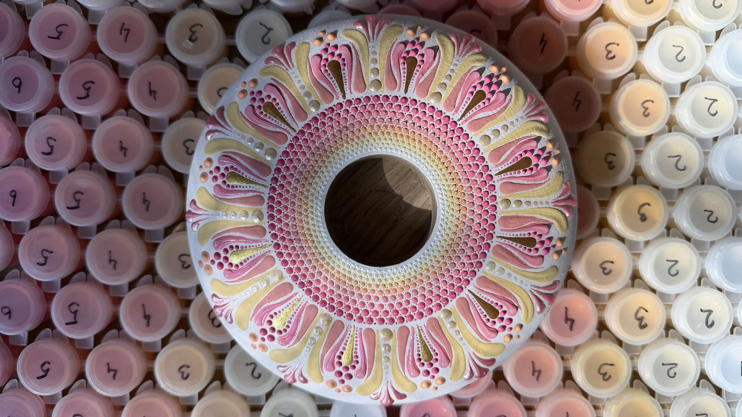 A circular object with intricate pink, gold, and white decorative patterns on a white background, surrounded by numerous small containers labeled with the numbers 1 through 9, arranged in a grid formation.