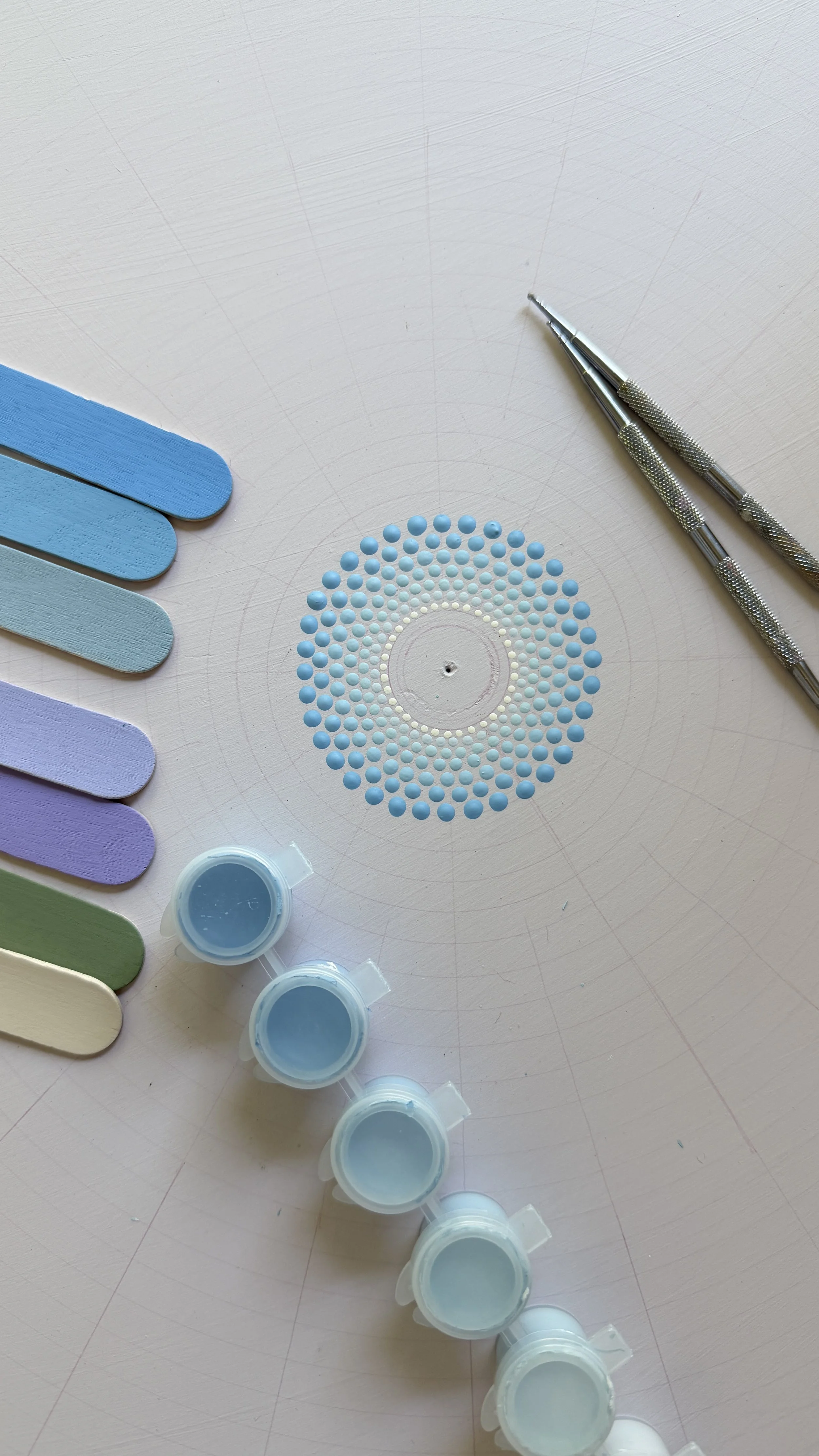 A palette of pastel nail polish colors, two metal dotting tools, and tiny containers of blue nail polish powder on a white circular surface.