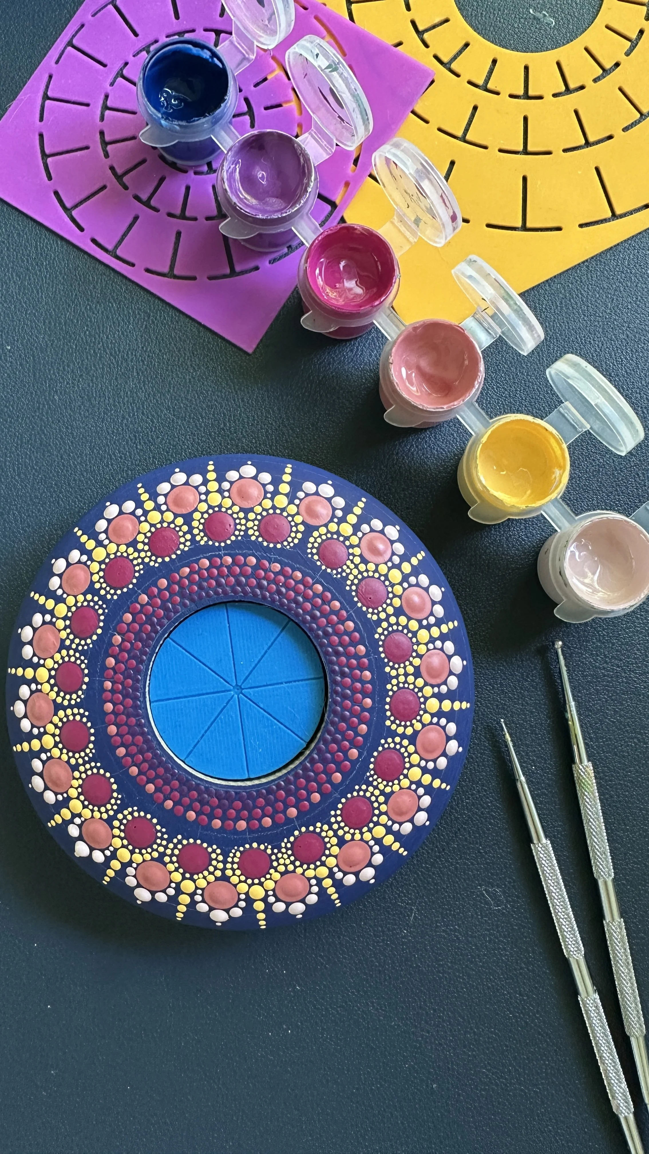 Nail art tools and paints, including a circular paint palette with pink and yellow dots, small pots of blue, purple, pink, and yellow paint, and two dotting tools on a black surface with colorful silicone nail stamping mats in pink, yellow, and purpl