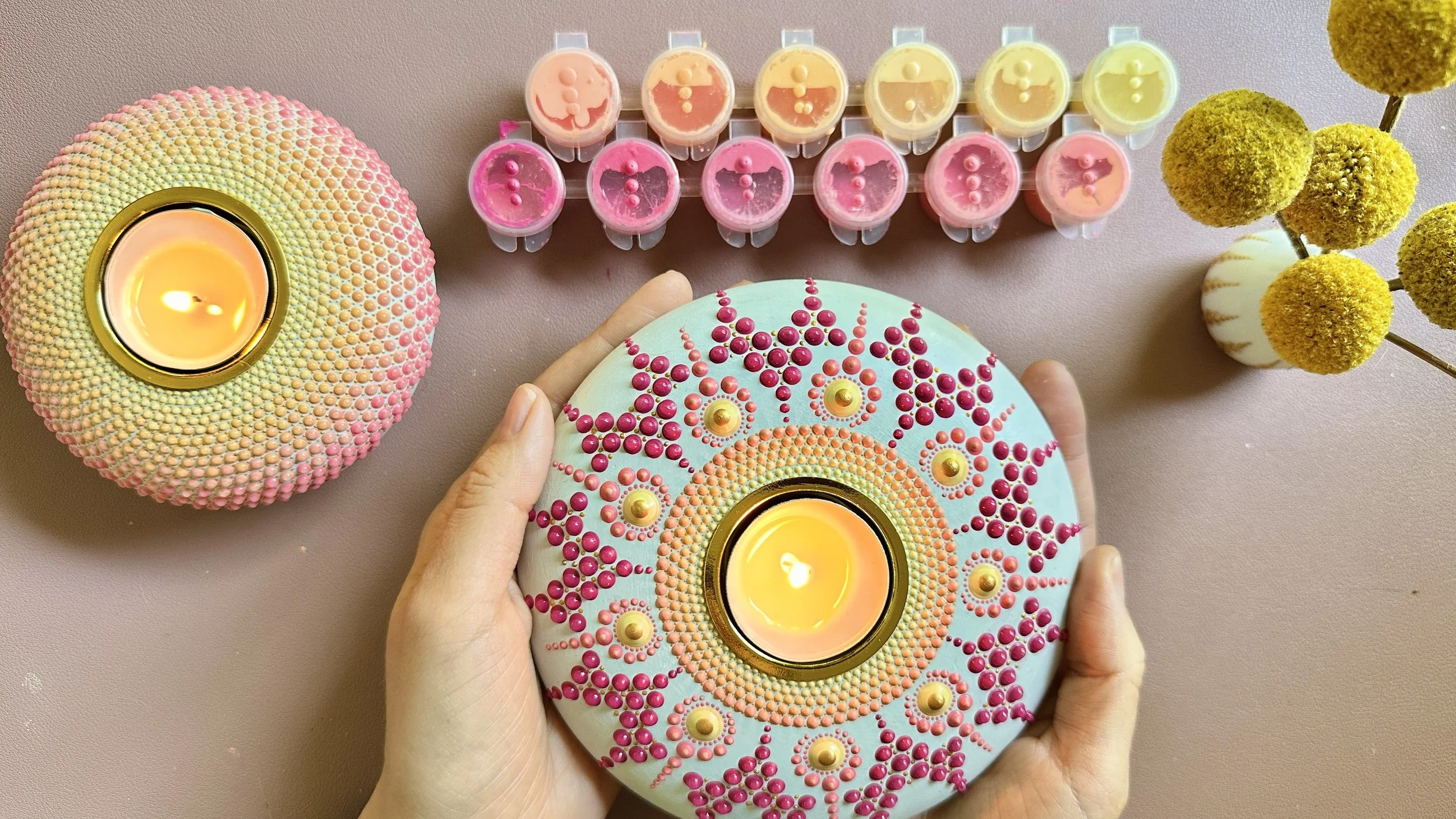 Decorative candle holder with intricate dot patterns and a lit candle inside, surrounded by small round flower arrangements and a row of pastel-colored lip balms.