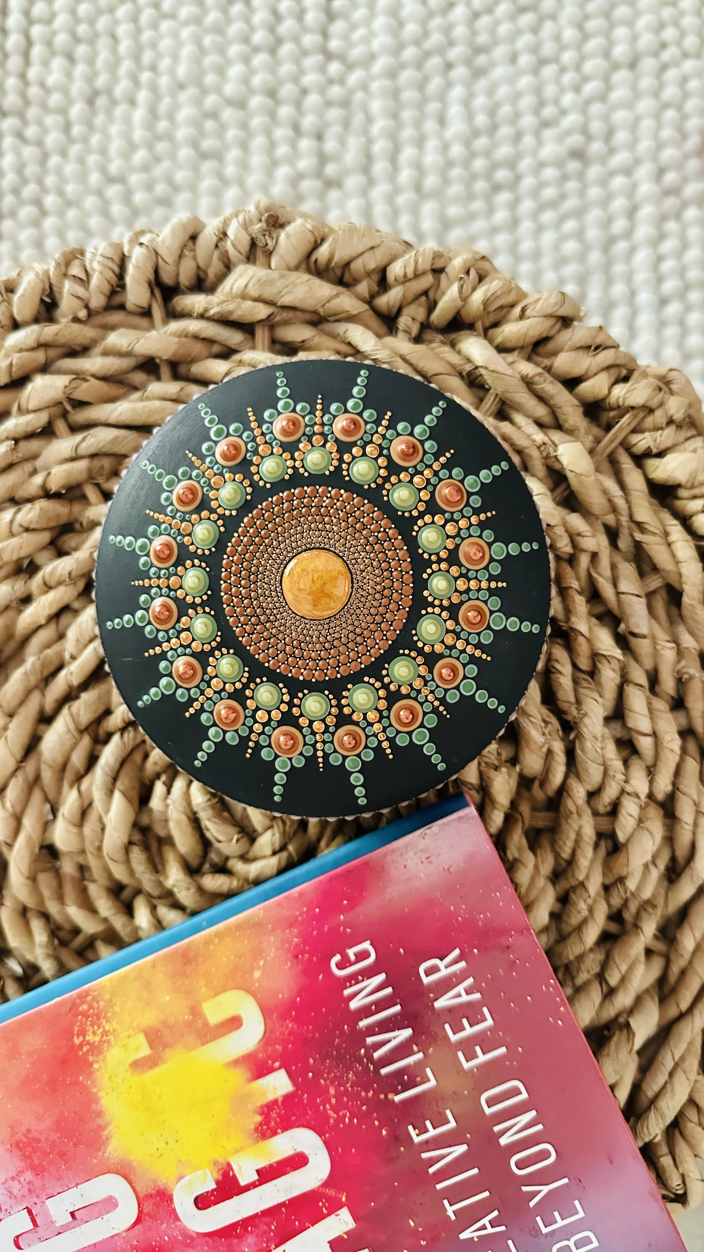A basket with a decorative circular item featuring a design with small colorful dots, placed on a textured woven surface, with part of a colorful book or magazine visible at the bottom.