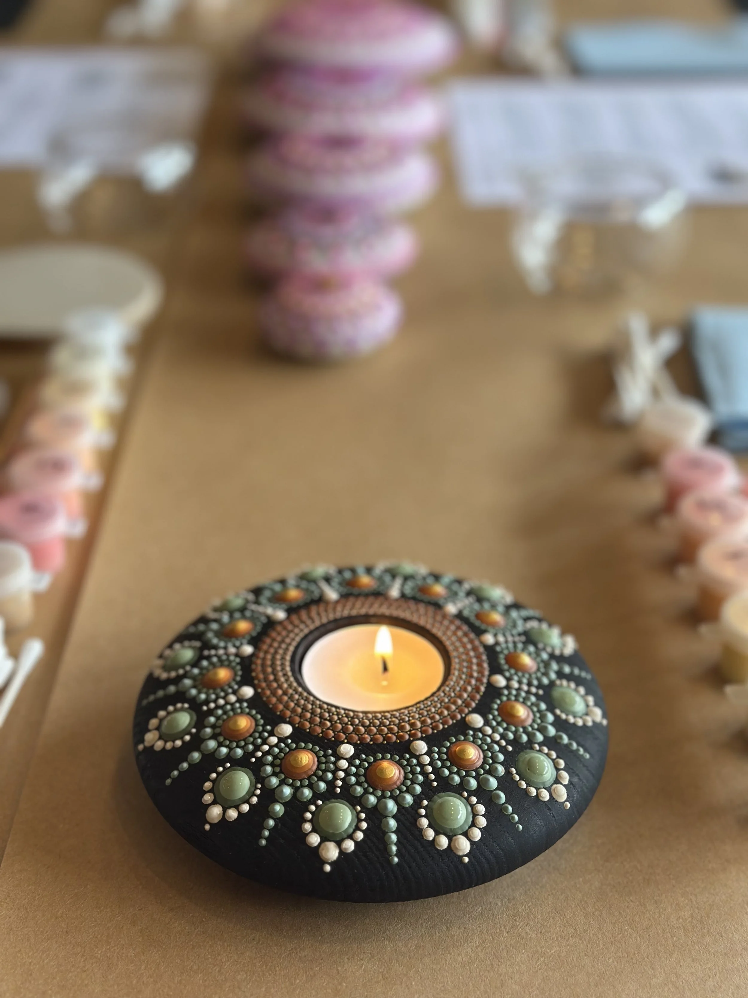 A painted black rock decorated with colorful dots surrounds a lit candle in a small dish, set on a flat surface.