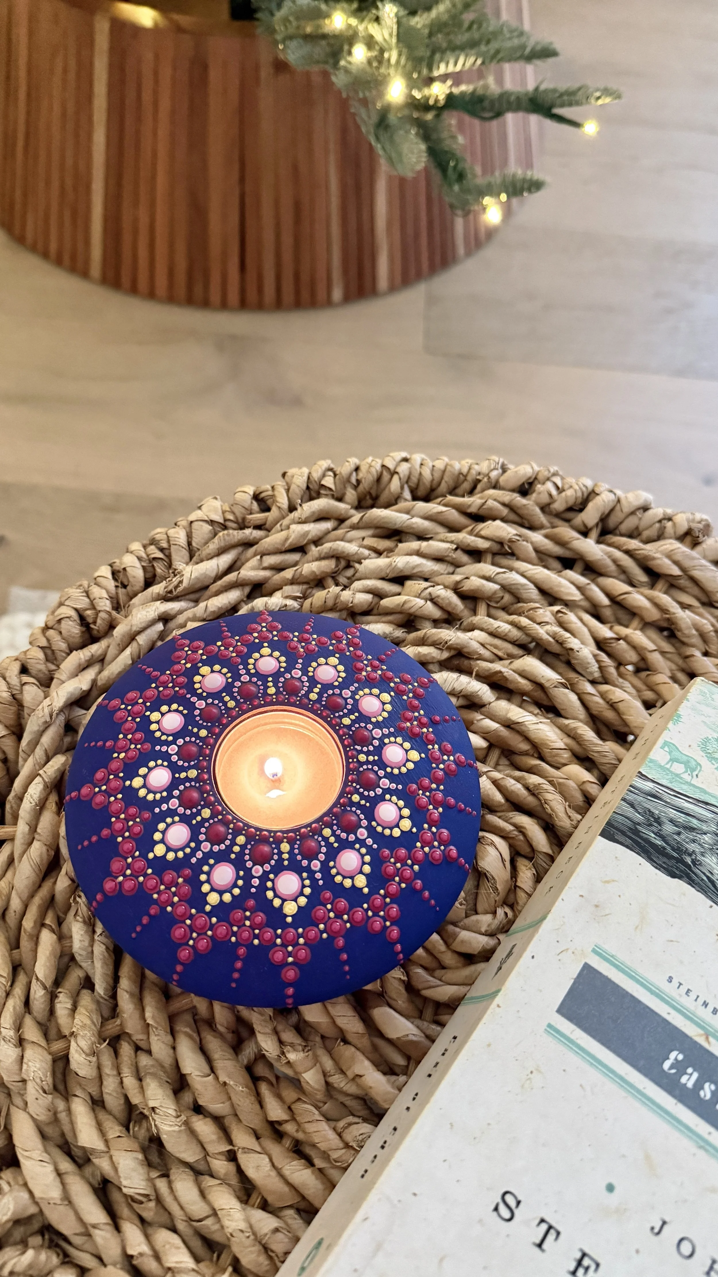 A painted candle holder with red and yellow dots forming a radial pattern, placed on a woven basket near a holiday card. In the background, a small lit Christmas tree is visible.