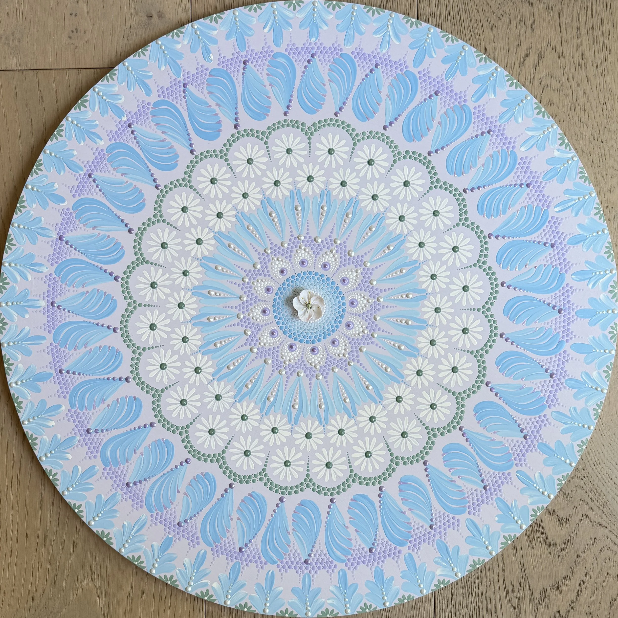 Decorative circular artwork with pastel blue, purple, white, and green colors, featuring leaf and floral motifs, placed on a wooden surface.