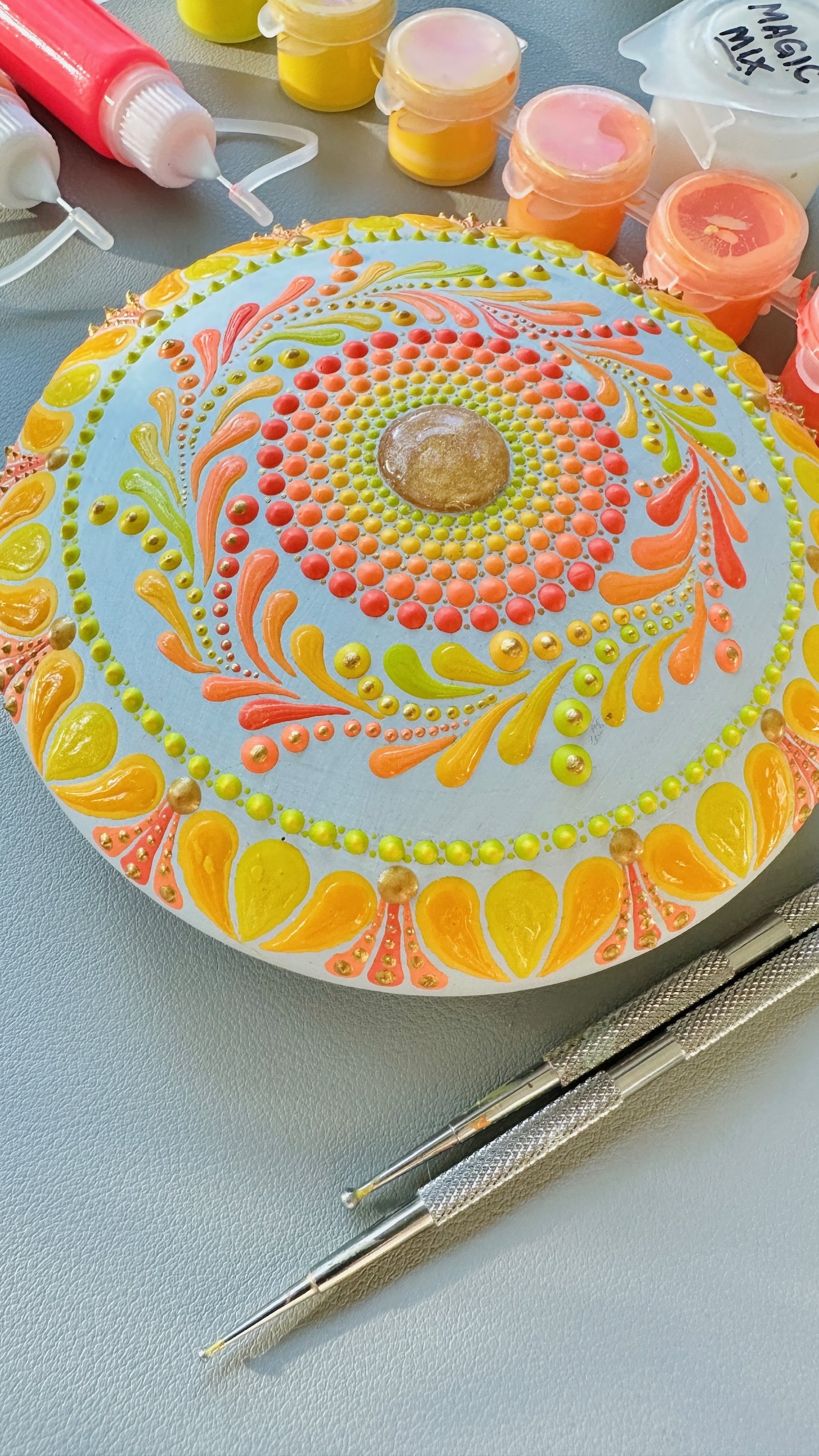 Colorful dot painting on a circular canvas with yellow, orange, pink, and green patterns, surrounded by skincare or cosmetic containers and tools on a blue surface.