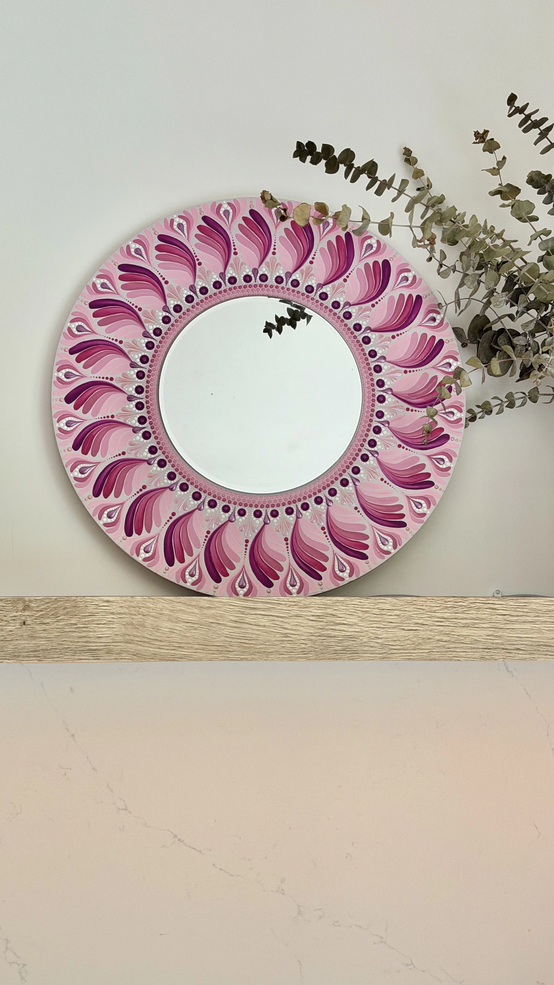 Decorative round mirror with a pink and purple feather pattern border, hanging on a white wall with dried eucalyptus branches nearby.