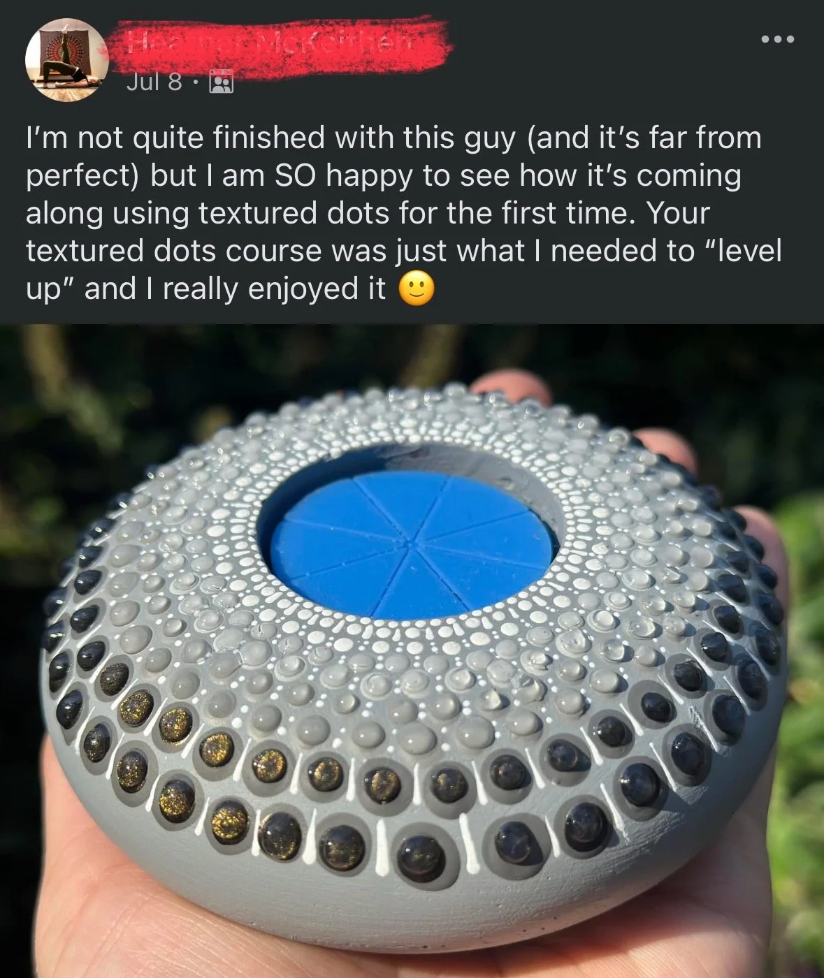 A close-up of a hand holding a spherical object with a textured surface covered in small black, gold, and gray dots. The object features a blue circular section with six segments in the center.