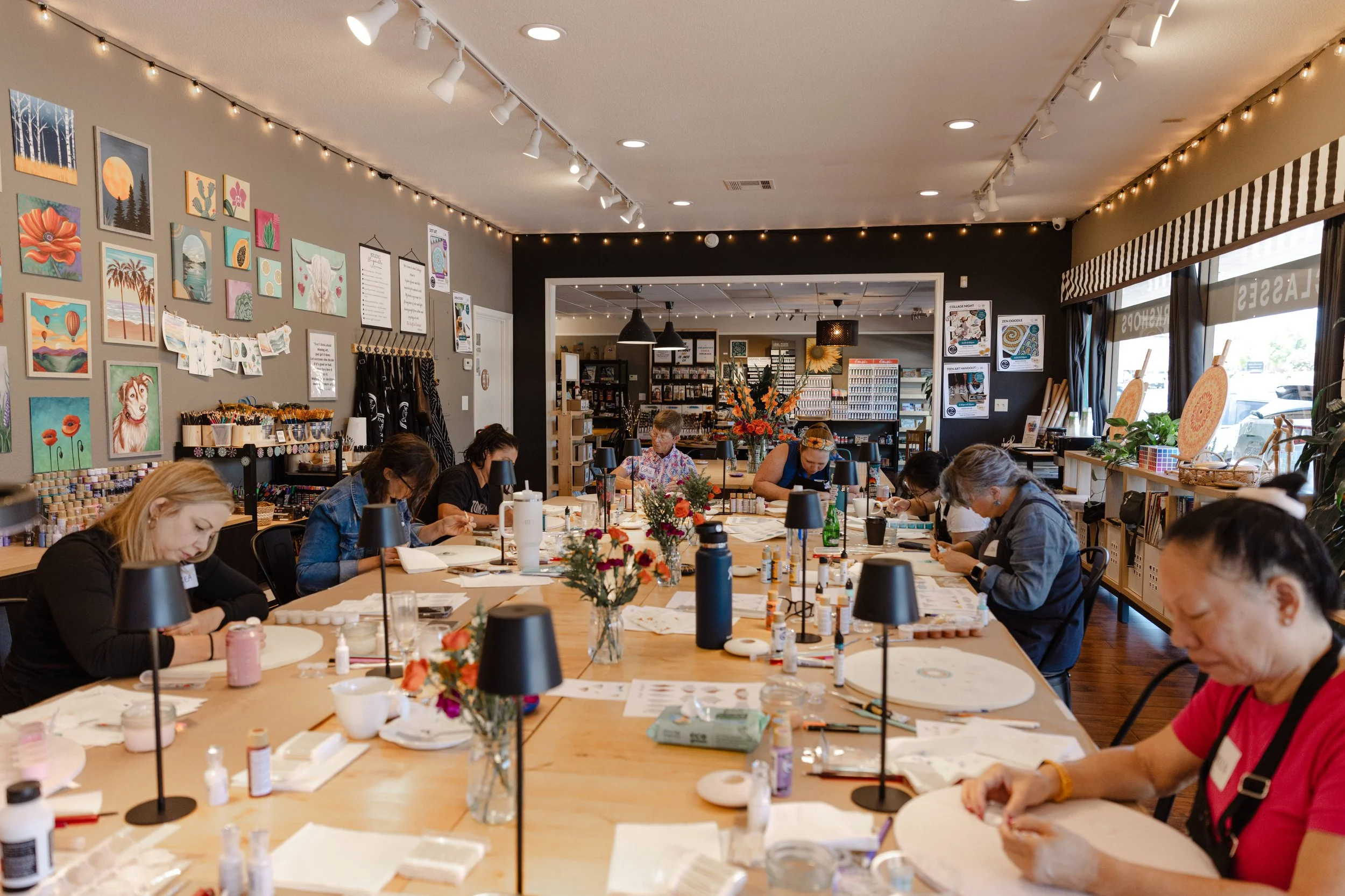 People are gathered around a large table in a well-lit room, engaging in an arts and crafts workshop. The room is decorated with framed artwork, string lights, and shelves filled with supplies.