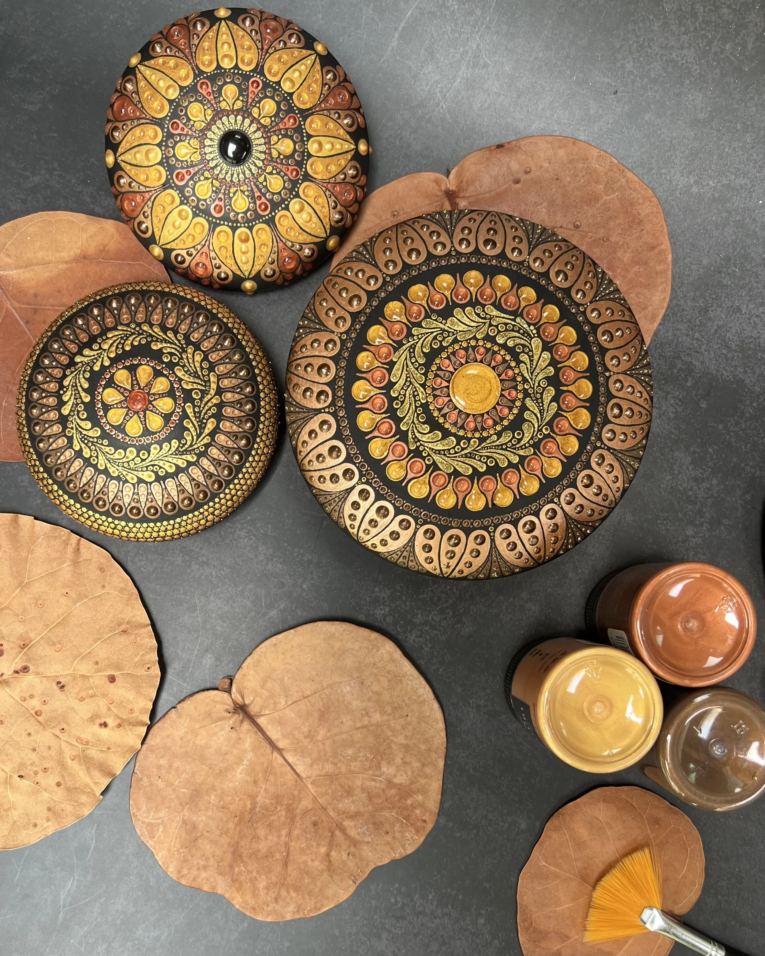 Decorative painted rocks with intricate patterns and colors, surrounded by dried leaves, and some jars of paint and a small fan-shaped brush on a dark surface.