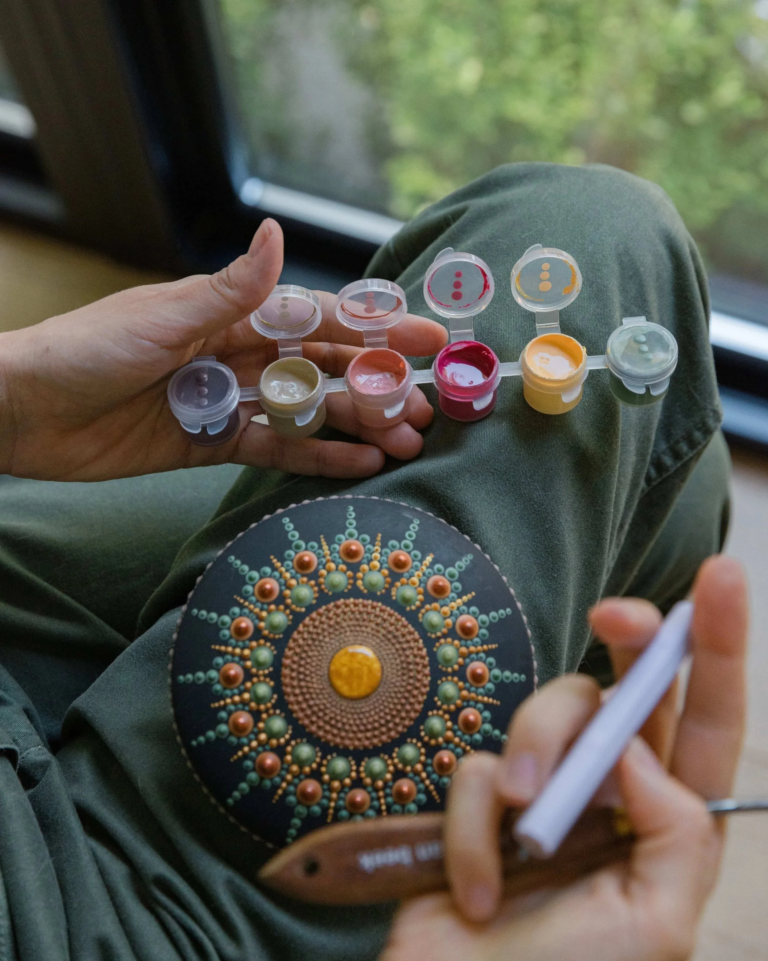 Person holding a palette of face paints and a container of dotting tools, while sitting on a bench near a window with green foliage outside.