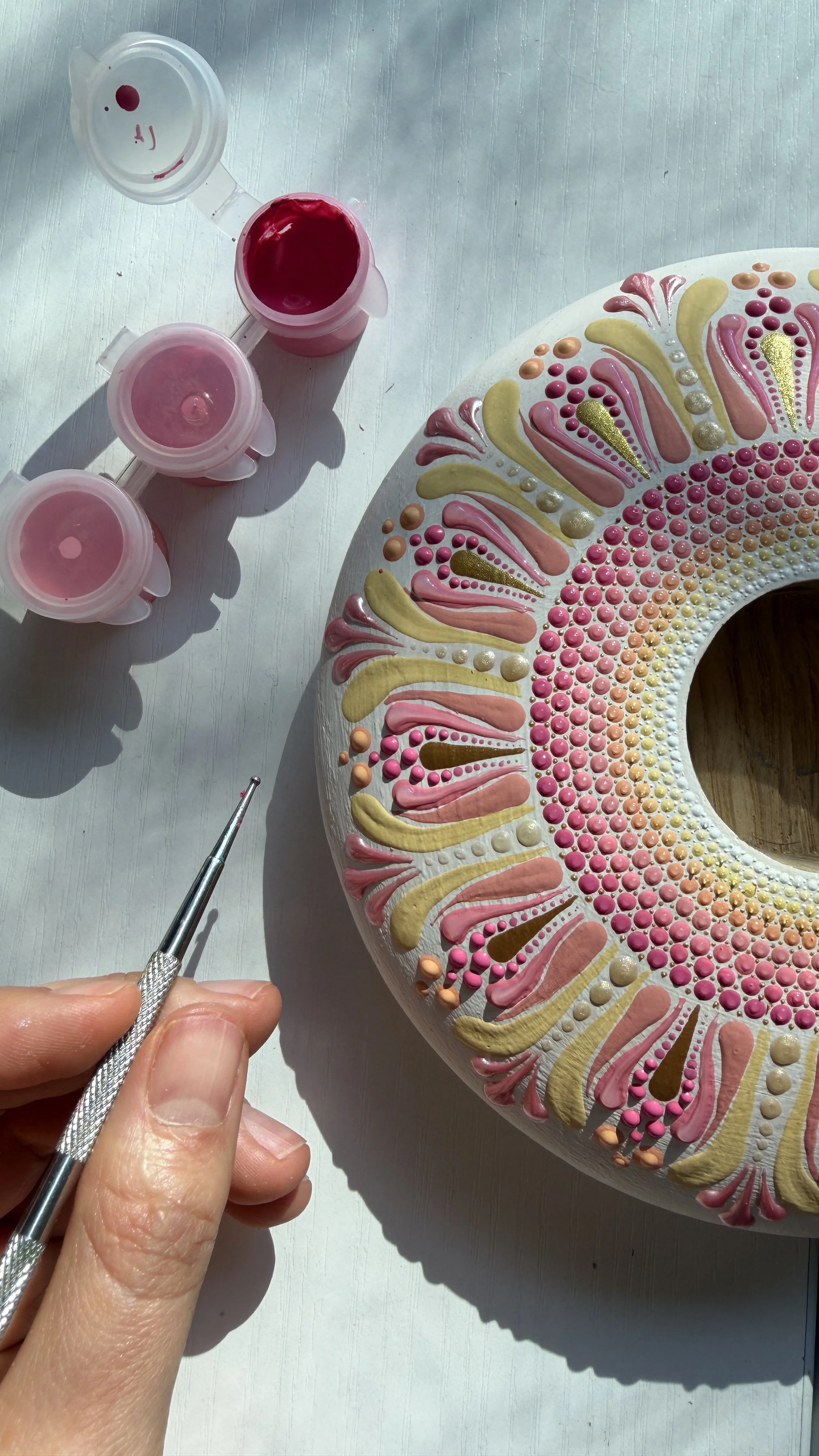 Person painting a decorative circular wooden object with pink, yellow, and white patterns using a fine brush, with three small containers of pink paint nearby.