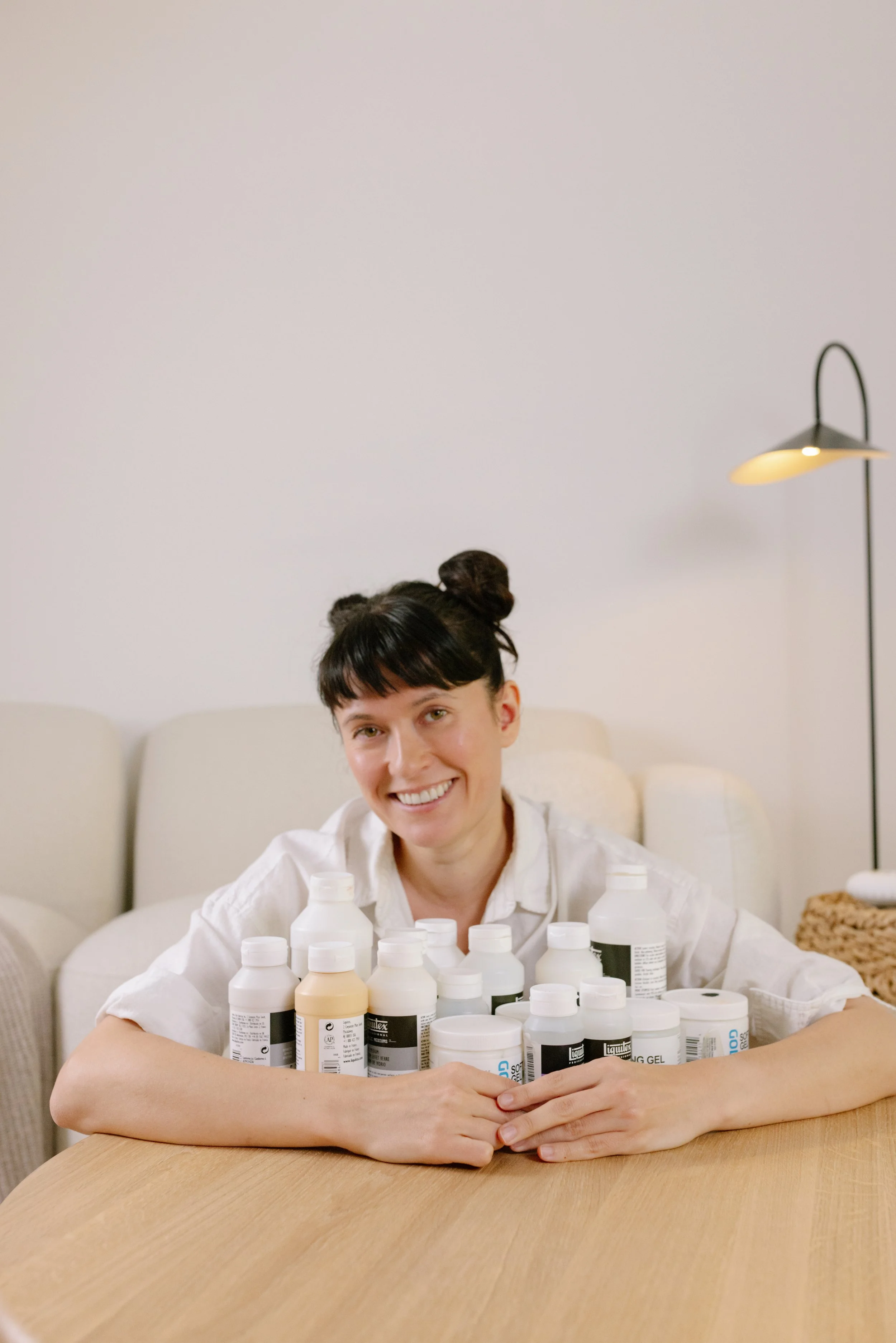 A woman with black hair styled in two buns, smiling, sitting at a wooden table surrounded by numerous bottles of skincare or cosmetic products, in a room with a white wall, a beige couch, and a floor lamp.