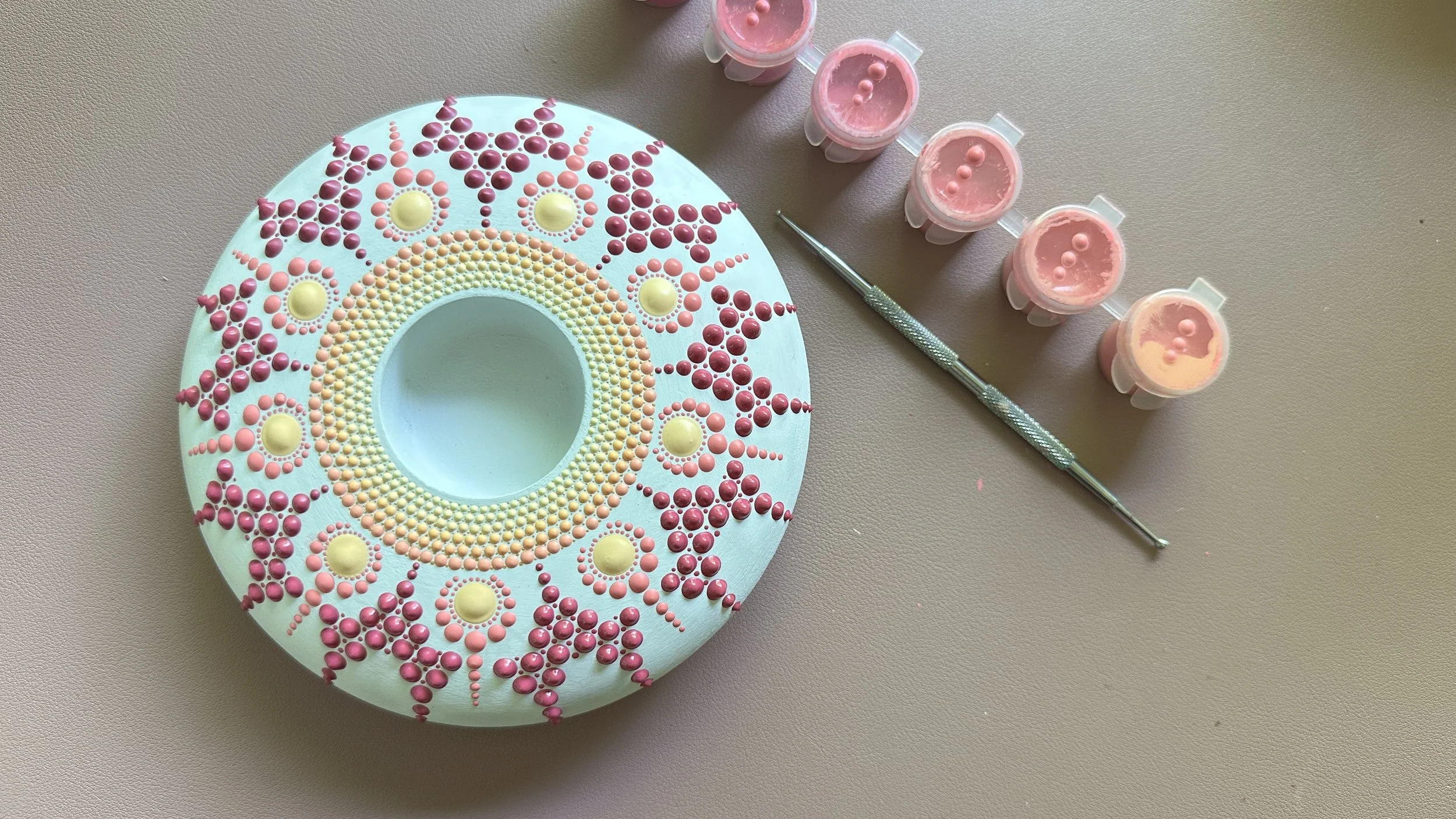 A decorative cake with pink and yellow candy dots arranged in a pattern, surrounded by four pink containers of icing and a small metal tool for decorating, all placed on a neutral-colored surface.