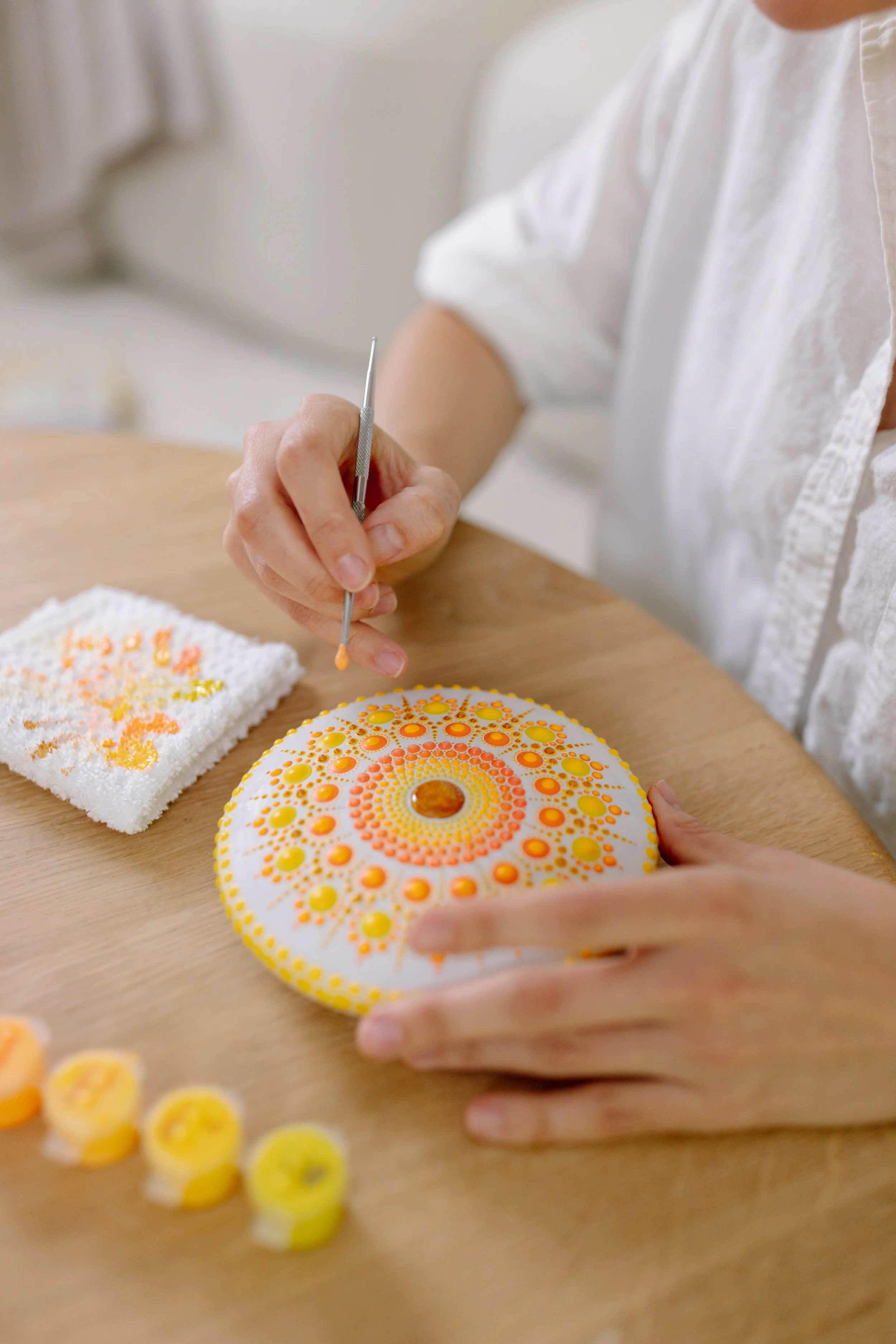 A person decorating a round, white cake with yellow and orange dot patterns using a small tool, with other decorated items and dots of icing nearby.