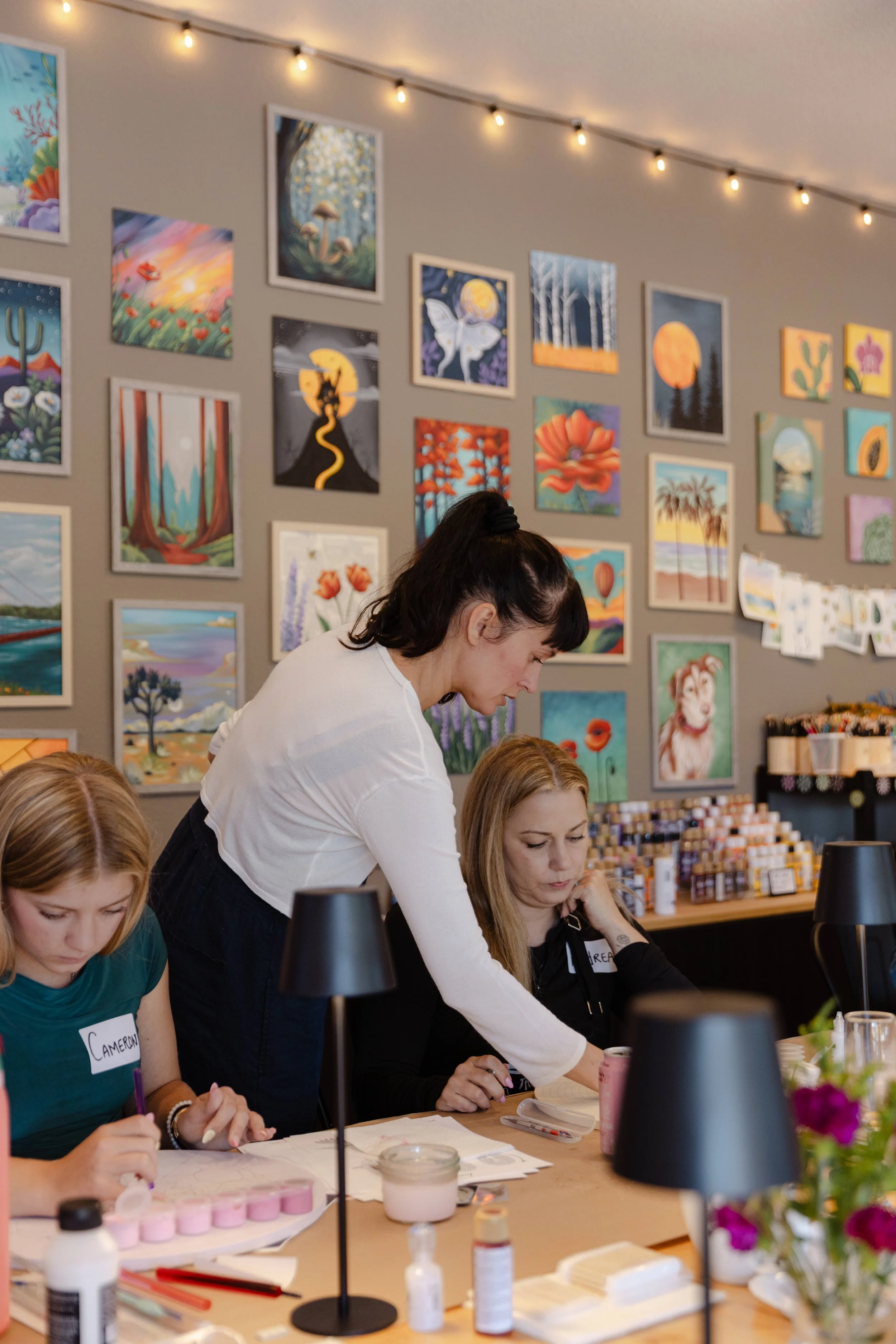 People gathered at a table involved in crafting or a workshop, with artwork paintings on the wall behind them and string lights overhead.
