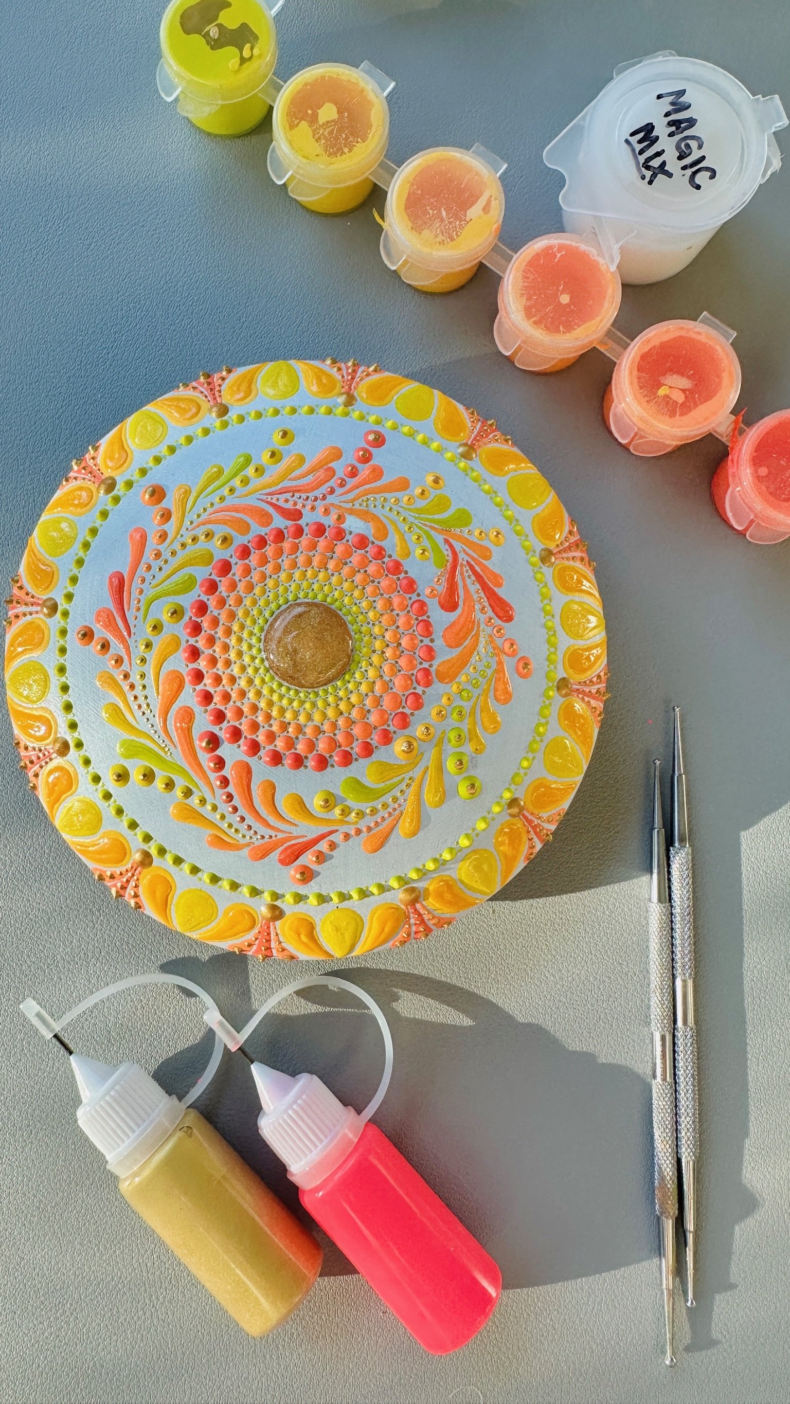 Colorful mandala cake decorated with pink, yellow, and orange icing dots and patterns, with a central brown candy. Surrounding the cake are tools and supplies for cake decorating, including squeeze bottles of yellow and pink icing, two precision deco