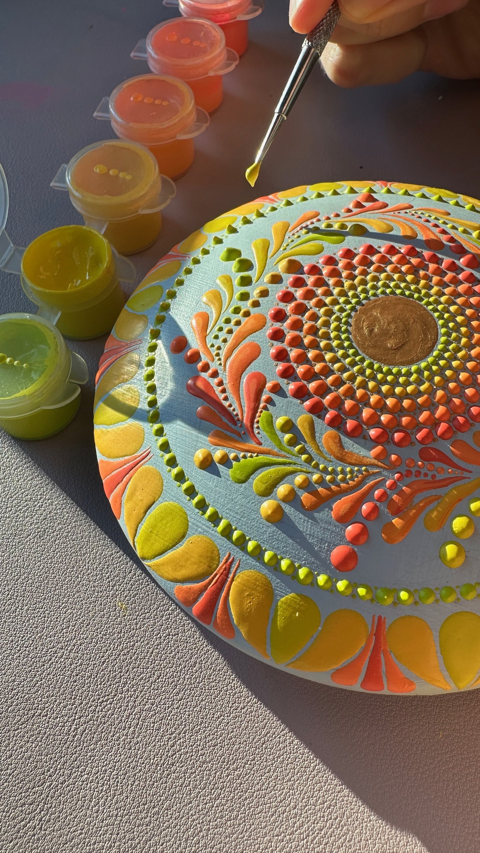 Colorful, detailed dot painting on a rounded canvas, with small containers of brightly colored paints nearby and a hand using a small tool to add detail to the artwork.