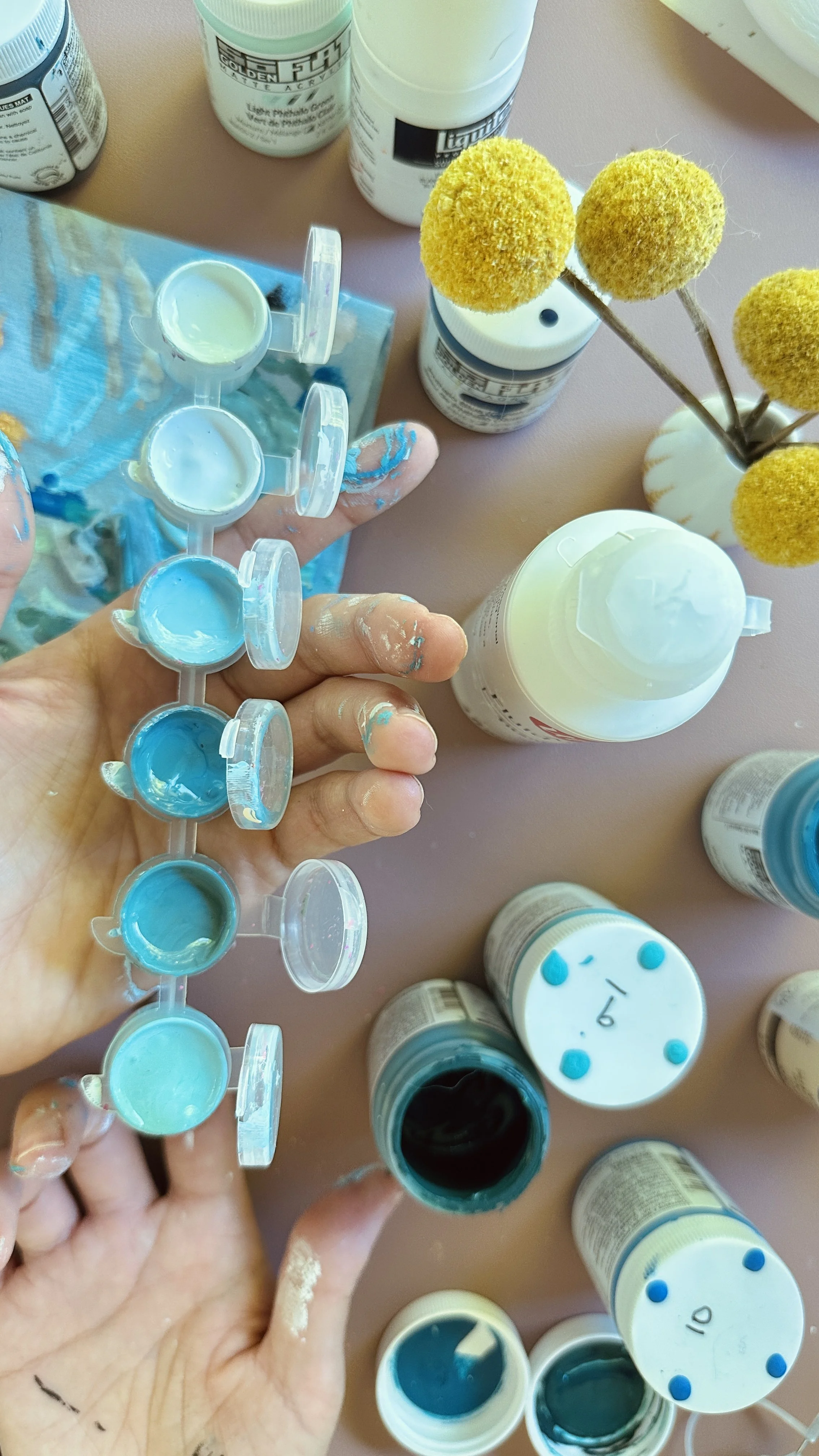 Person holding a paint palette with various shades of blue and teal, surrounded by bottles of paint and craft supplies, some partially spilled on their fingers.