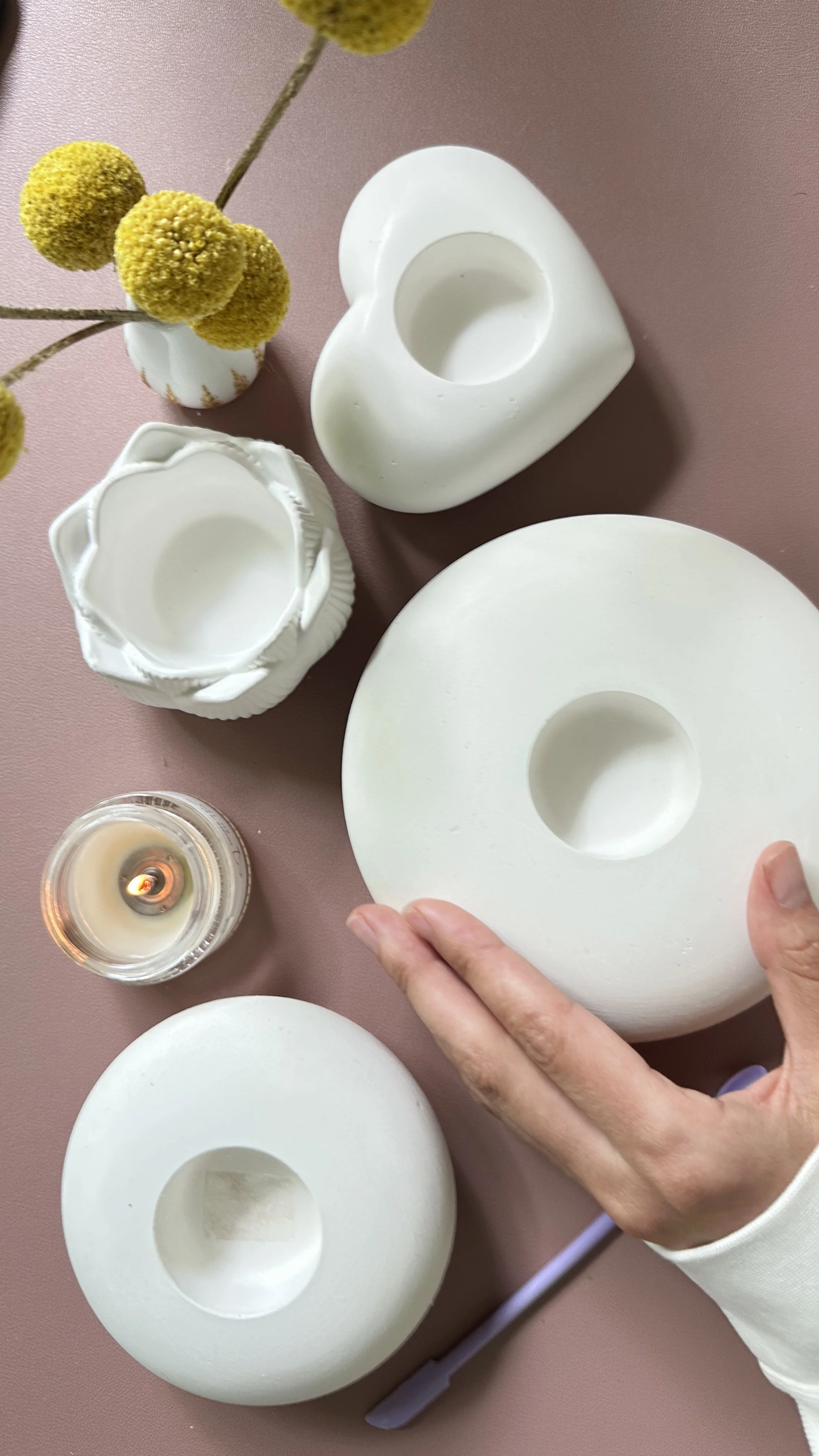 Person holding a white ceramic plate with hollow center, surrounded by similar plates, a lit candle, a vase with yellow pom-pom flowers, and a pink surface.