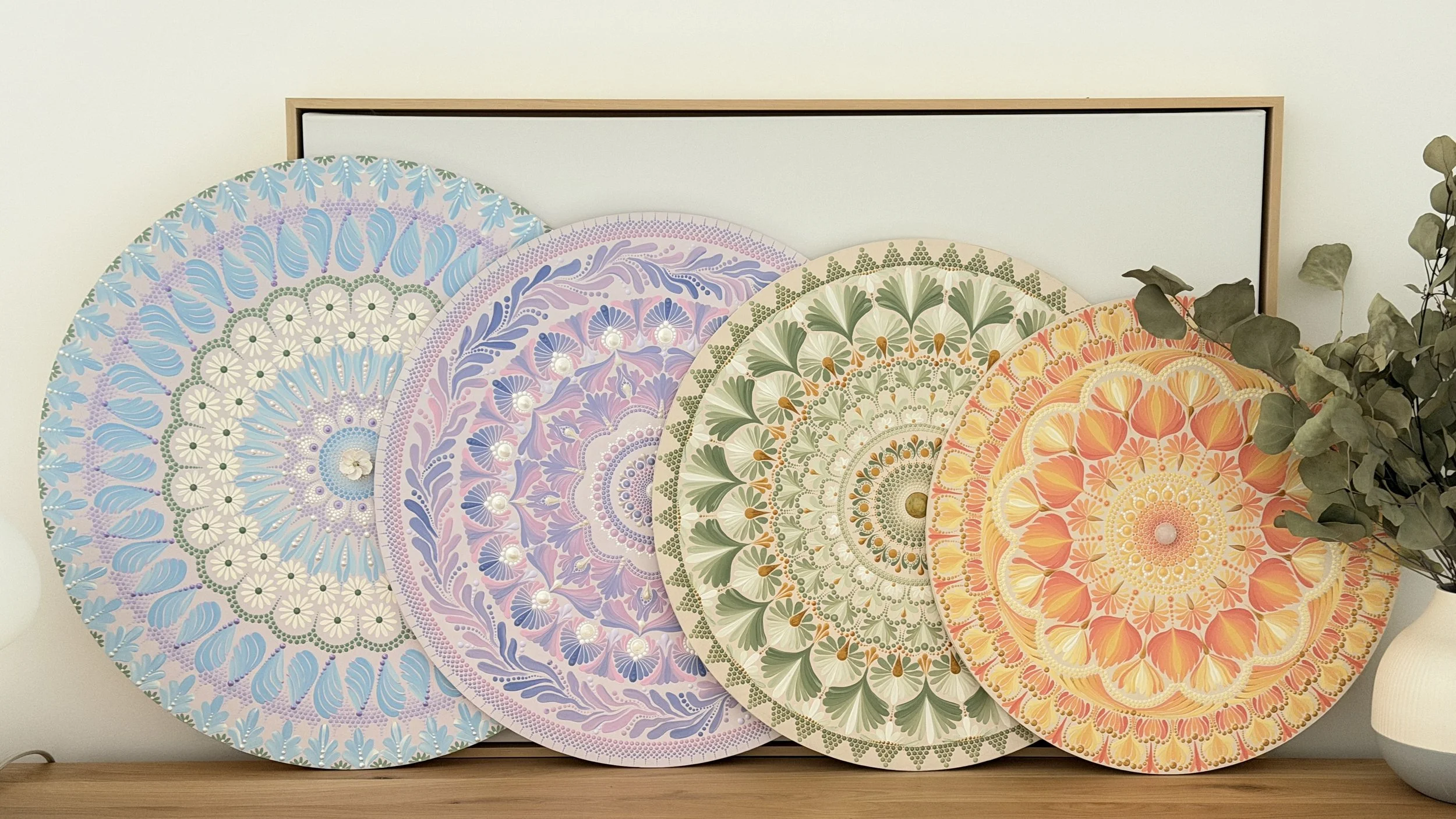 Five decorative circular mandala-style artworks with floral patterns in blue, purple, green, yellow, and orange, leaning against a wall with a small plant on the right.