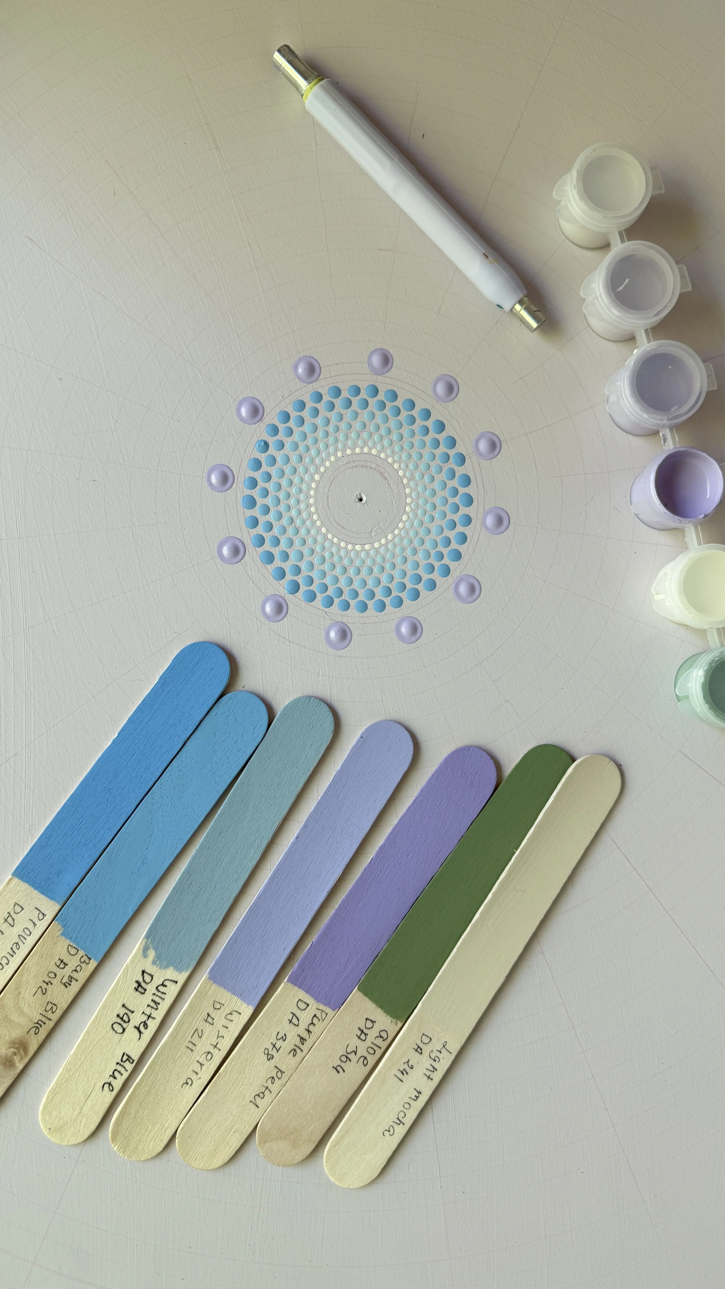 Color swatches labeled with various paint color names, arranged on a white surface along with small bottles of paint, a mixing stick, and a white pen.