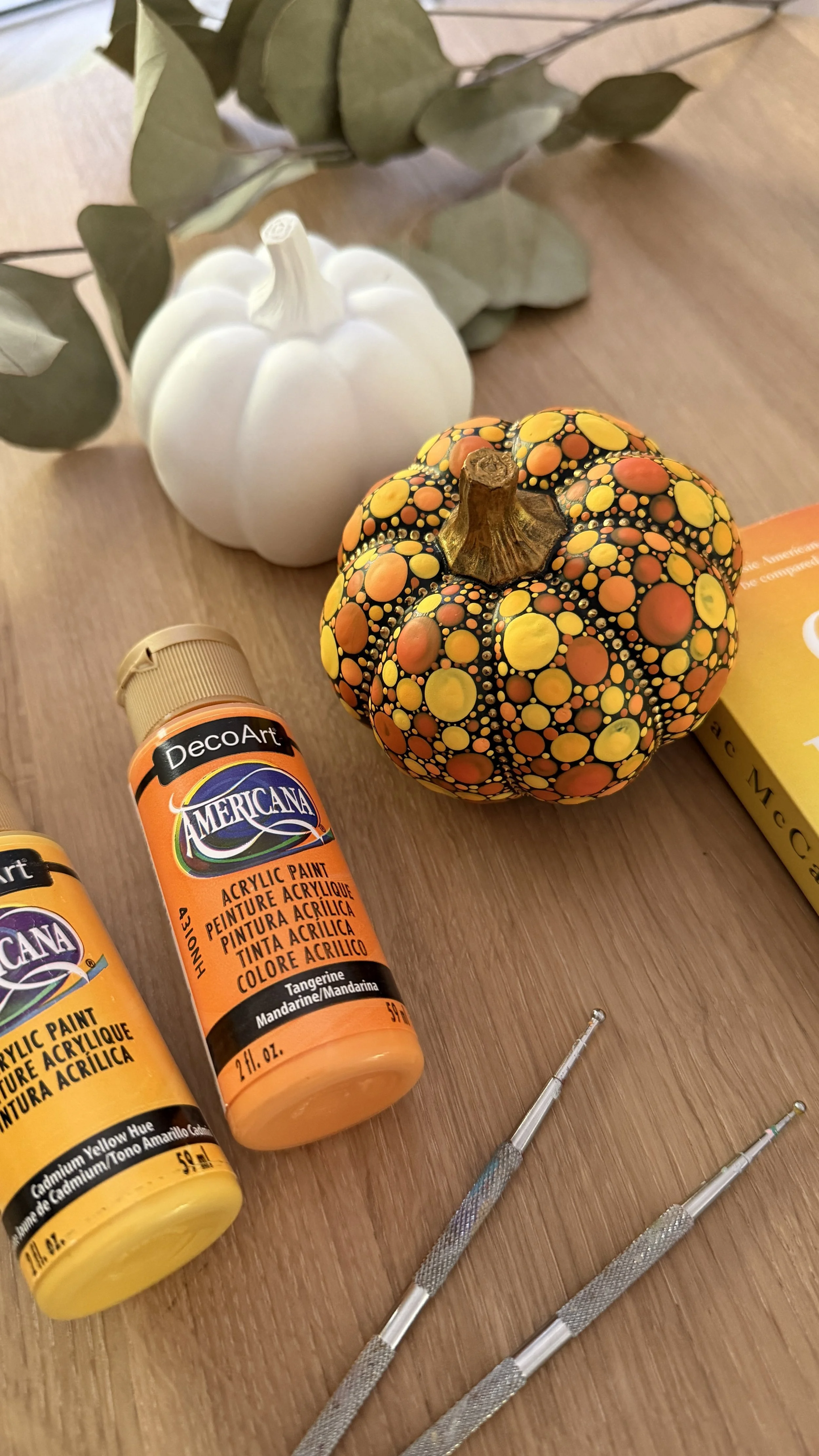 Decorative white pumpkin, painted black pumpkin with yellow, orange, and red circles, bottles of yellow and orange acrylic paint, and small precision screwdrivers on a wooden surface.