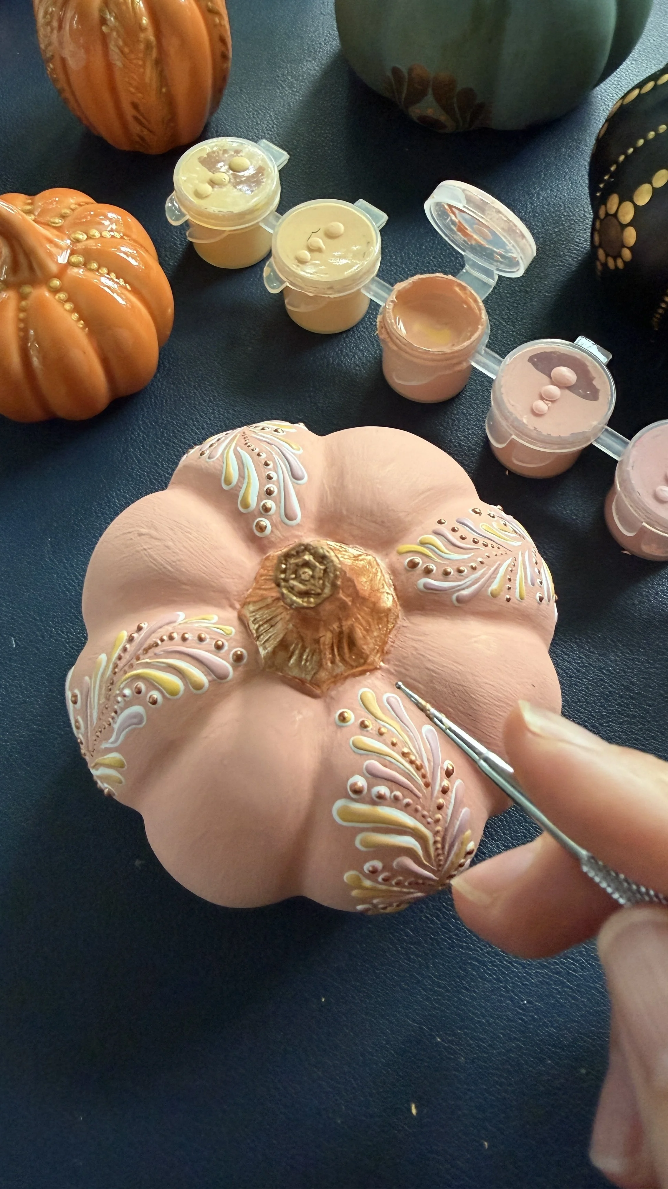 Hand painting intricate floral designs with paint on a decorative light pink pumpkin, surrounded by small pumpkins and gourds.