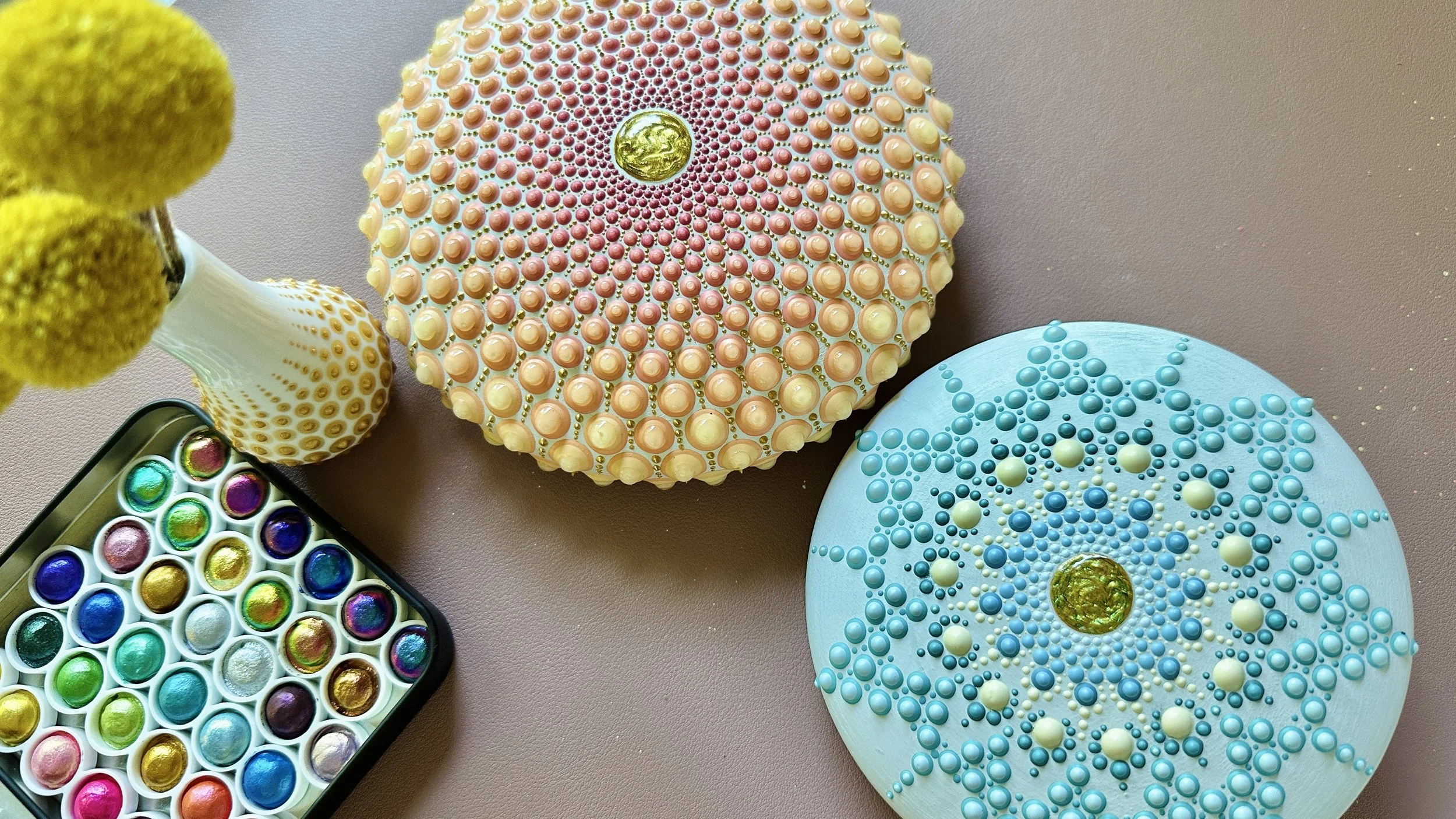 Close-up of colorful, patterned fidget toys on a pink surface, including two circular items with dotted designs and a rectangular container filled with multicolored gel beads.