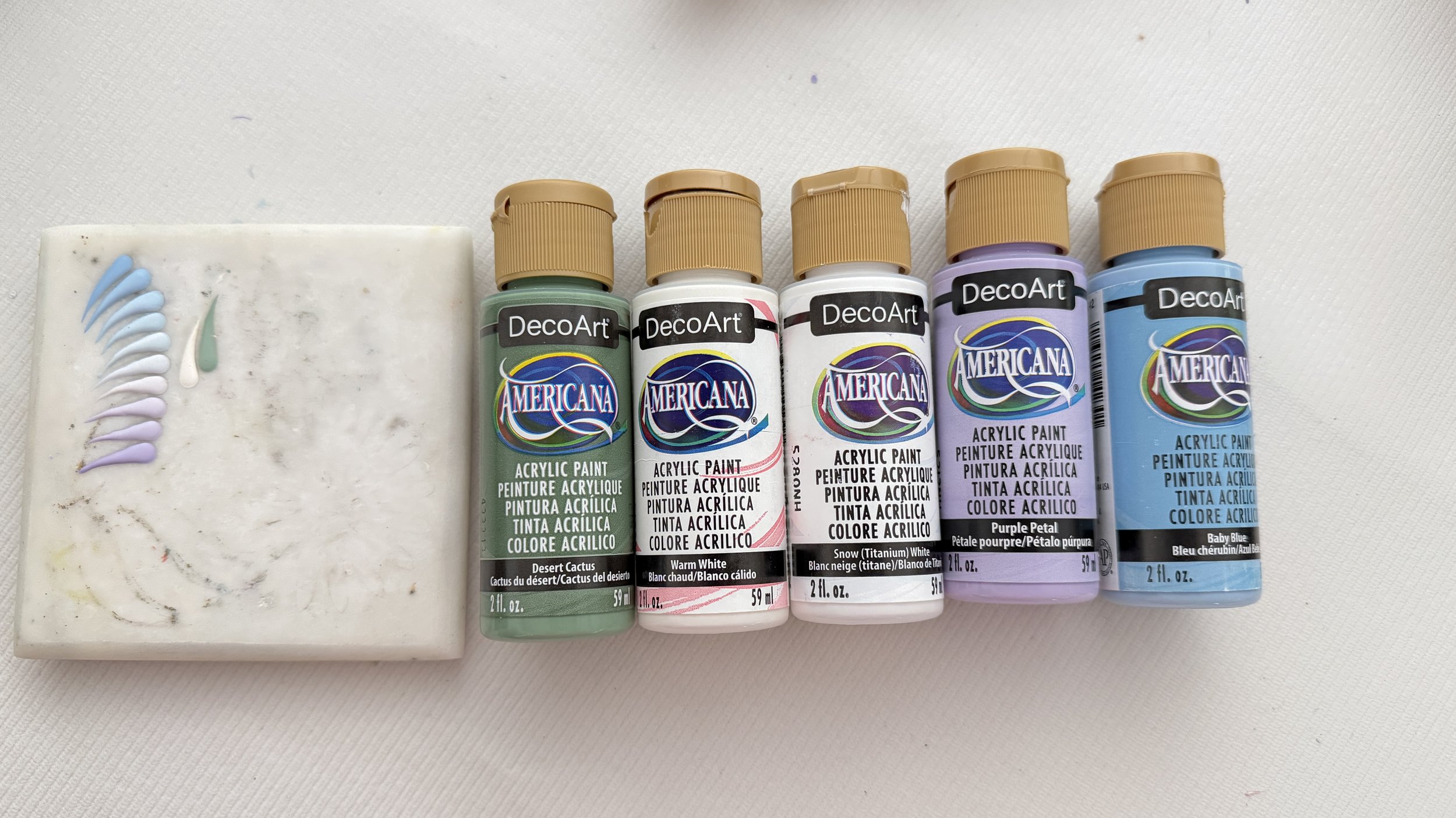 Five bottles of Americana DecoArt acrylic paint in various colors including cactus, white, snow, purple petal, and baby blue, placed in a row on a white surface with a paint sample on a white palette board to the left.