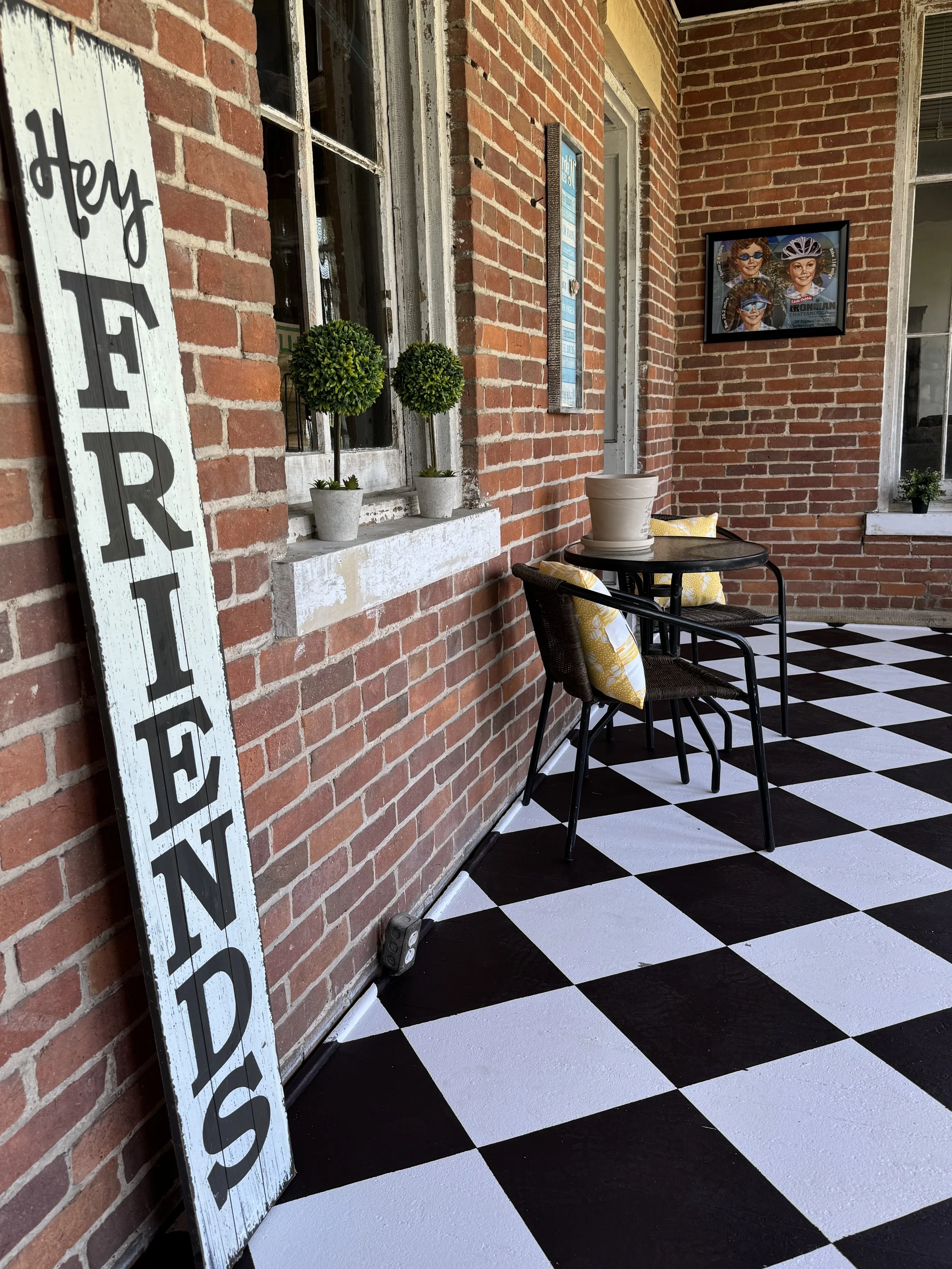 Porch with brick wall, decorative green plants in pots, a black table with two yellow patterned pillows, a large vertical white sign with black letters. The  painted floor has a black and white checkerboard pattern.