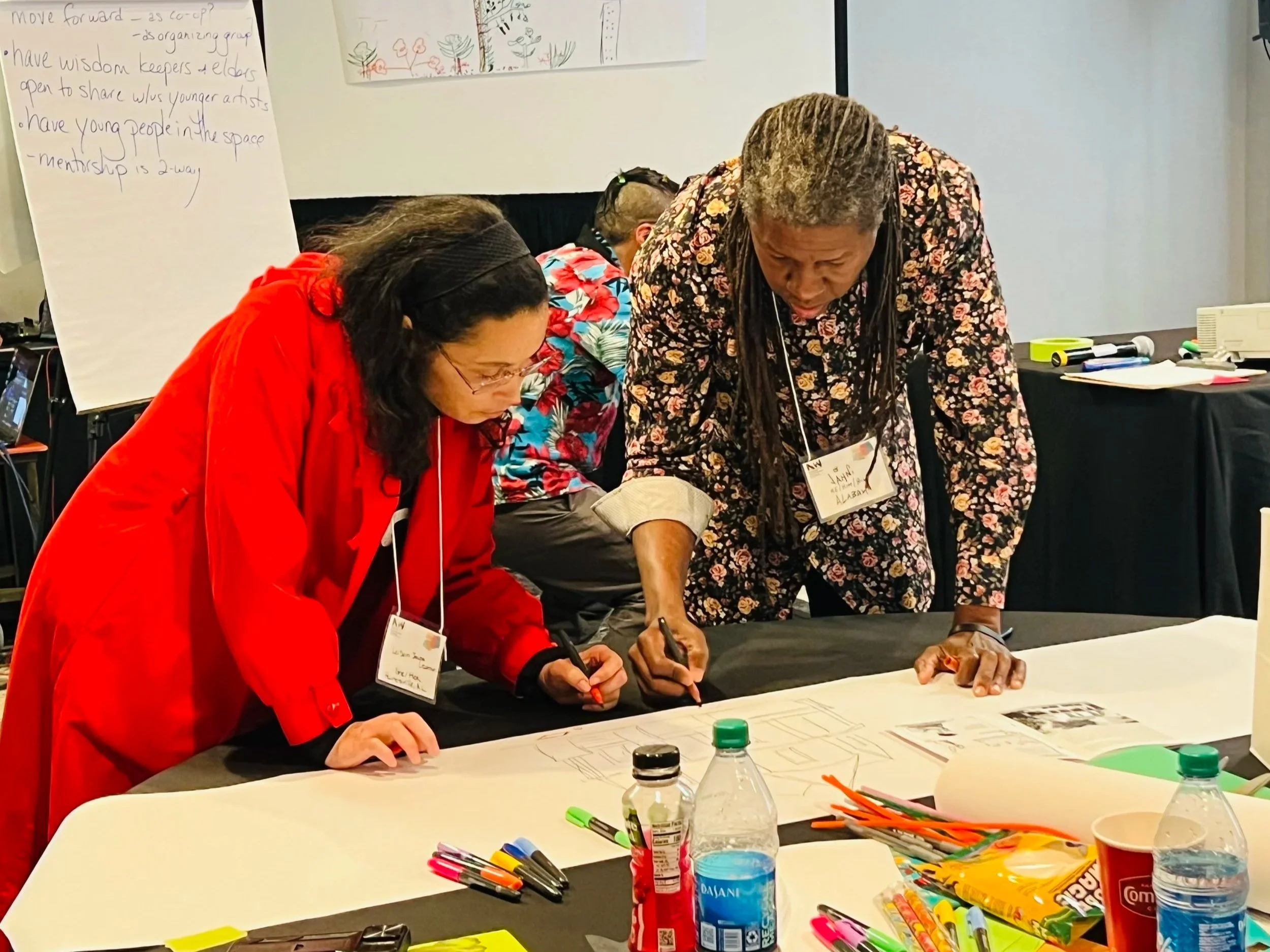 Two women are working together on paper drawings at a table during a workshop or conference. One woman wears a red jacket, and the other wears a floral shirt. The table has markers, water bottles, snacks, and other supplies.