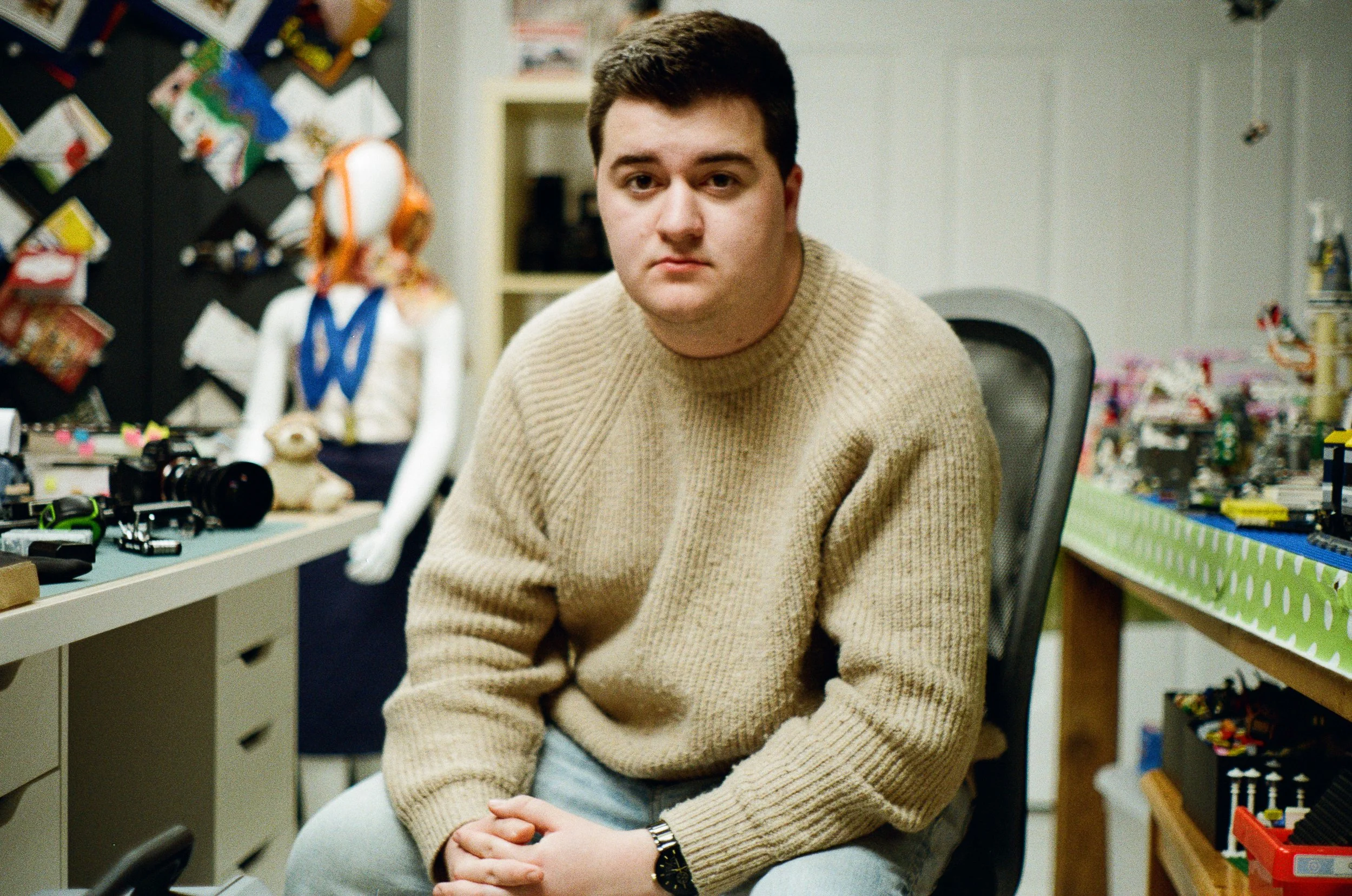A young man with dark hair, wearing a beige sweater, sitting in an office or workshop, with colorful items and toys around him, looking directly at the camera with a neutral expression.