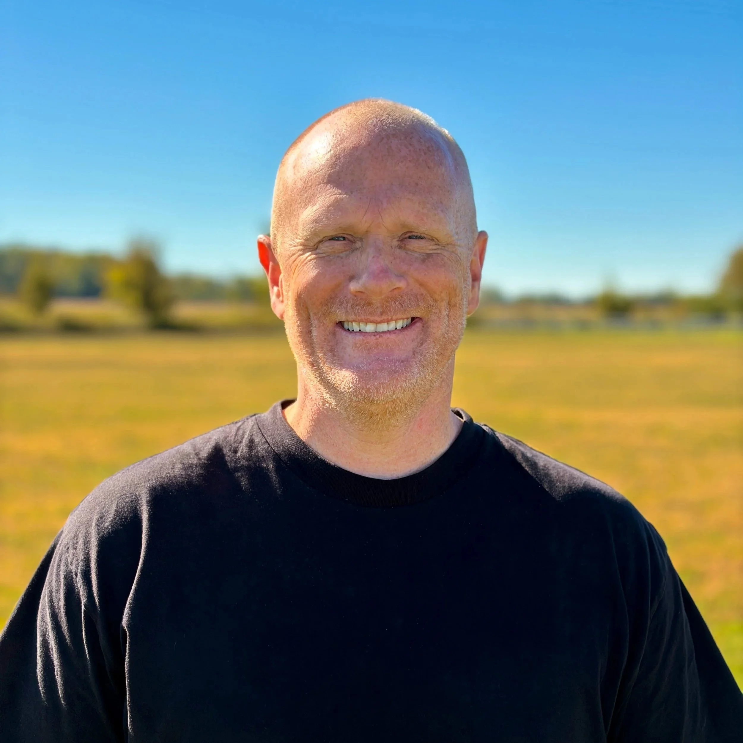 A smiling man with a red beard and bald head, wearing a navy blue t-shirt and light gray pants, standing in a room with beige walls, a brown couch, and a blurred green plant in the background.