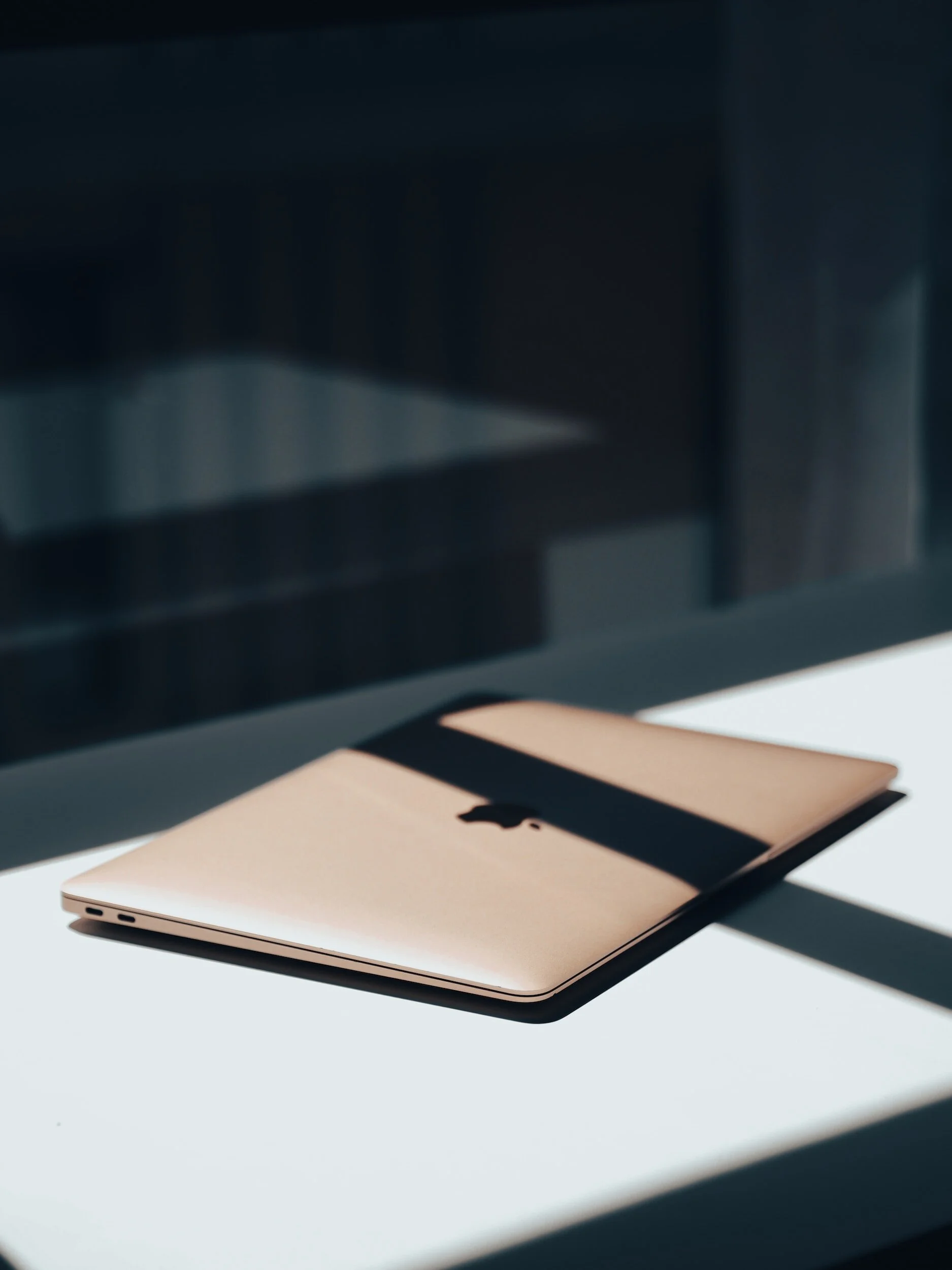 A closed gold-colored Apple MacBook laptop placed on a light-colored surface with shadow from nearby window or object.