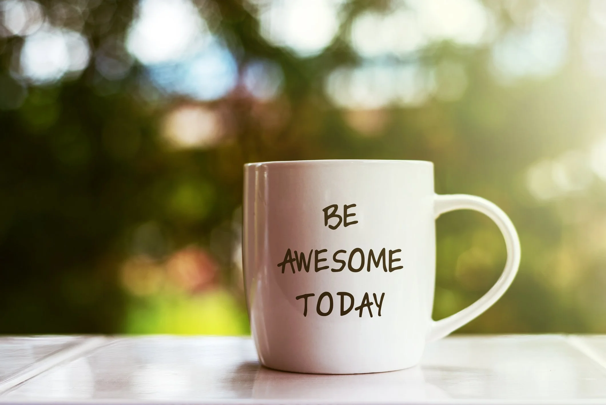 White coffee mug with the words 'BE AWESOME TODAY' printed on it, placed on a white surface with a blurred outdoor background.