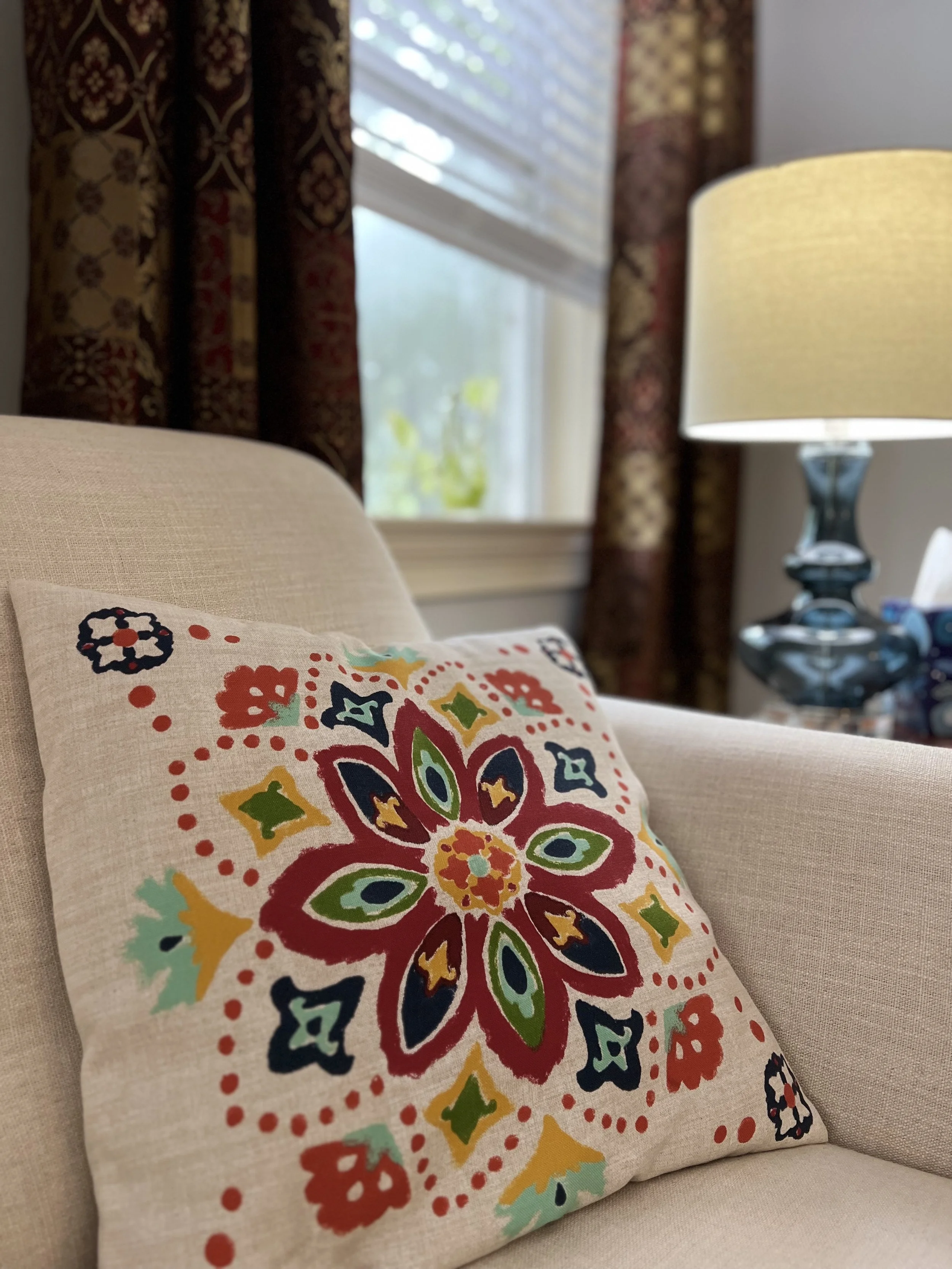 Close-up of a decorative pillow with a colorful floral pattern on a beige sofa at Heart Mind & Soul Counseling in Hilton Head Island, SC, with a window, curtains, and a table lamp in the background.