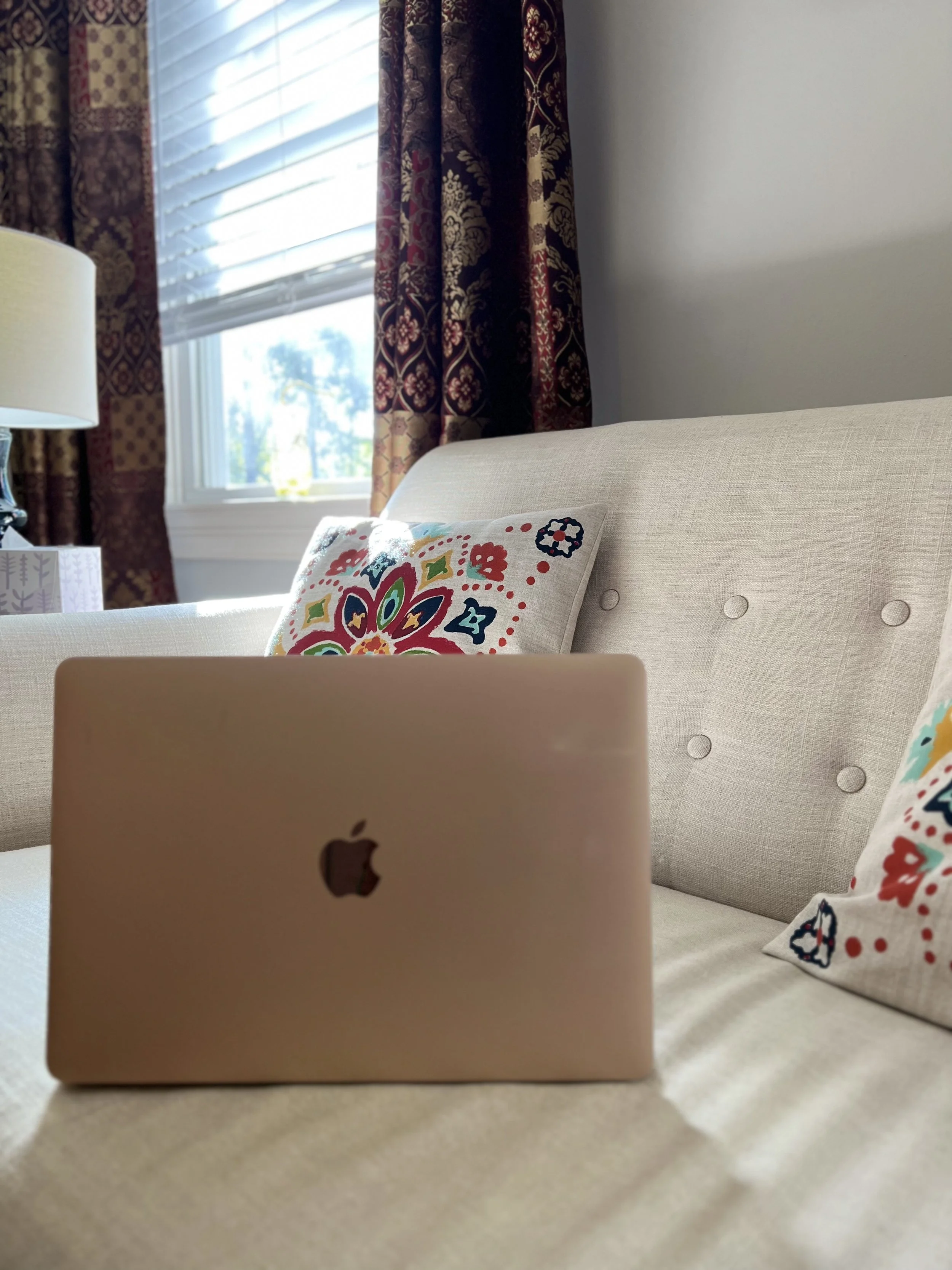 Close-up of gold laptop at Heart Mind & Soul Counseling in Hilton Head Island, SCon a beige couch with embroidered pillows in front of a window with patterned curtains and blinds, in a well-lit living room.