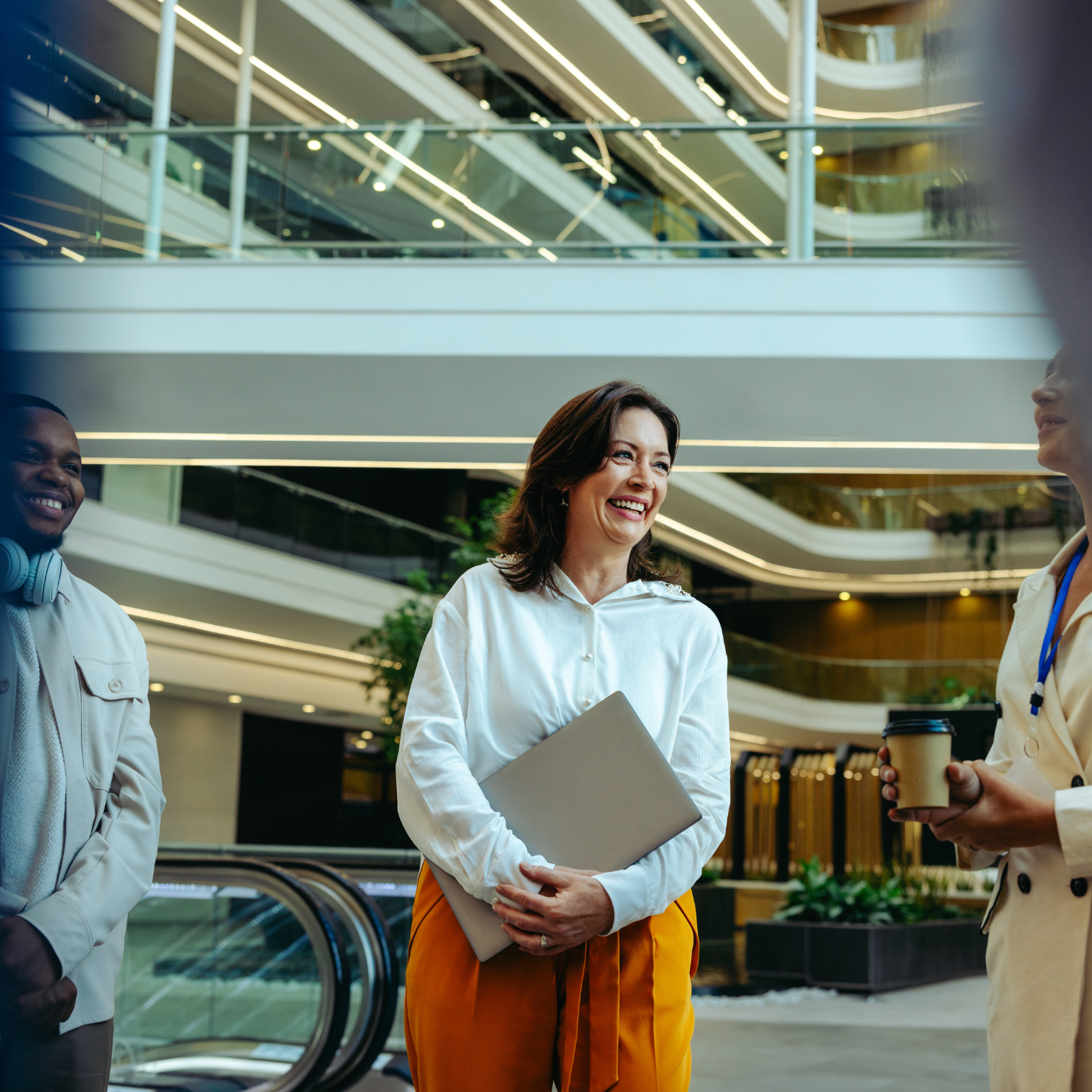 a middle aged latina woman stands in a workspace lobby holding her laptop while standing next to a young man of color wearing headphones, both are smiling as they look to a woman speaking to them